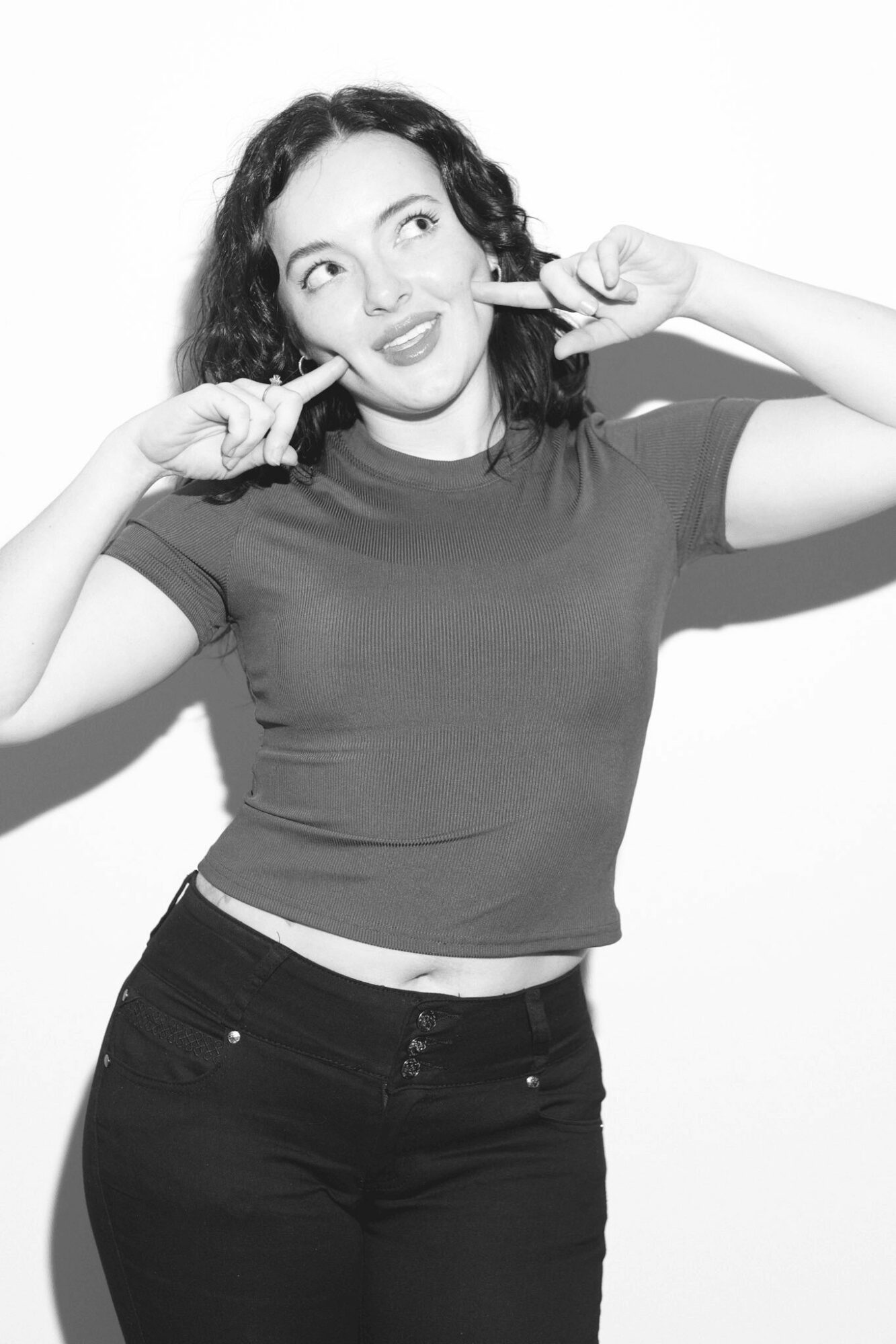 Young woman with curly hair making a playful face, touching her cheeks with fingers, wearing a fitted t-shirt and jeans.