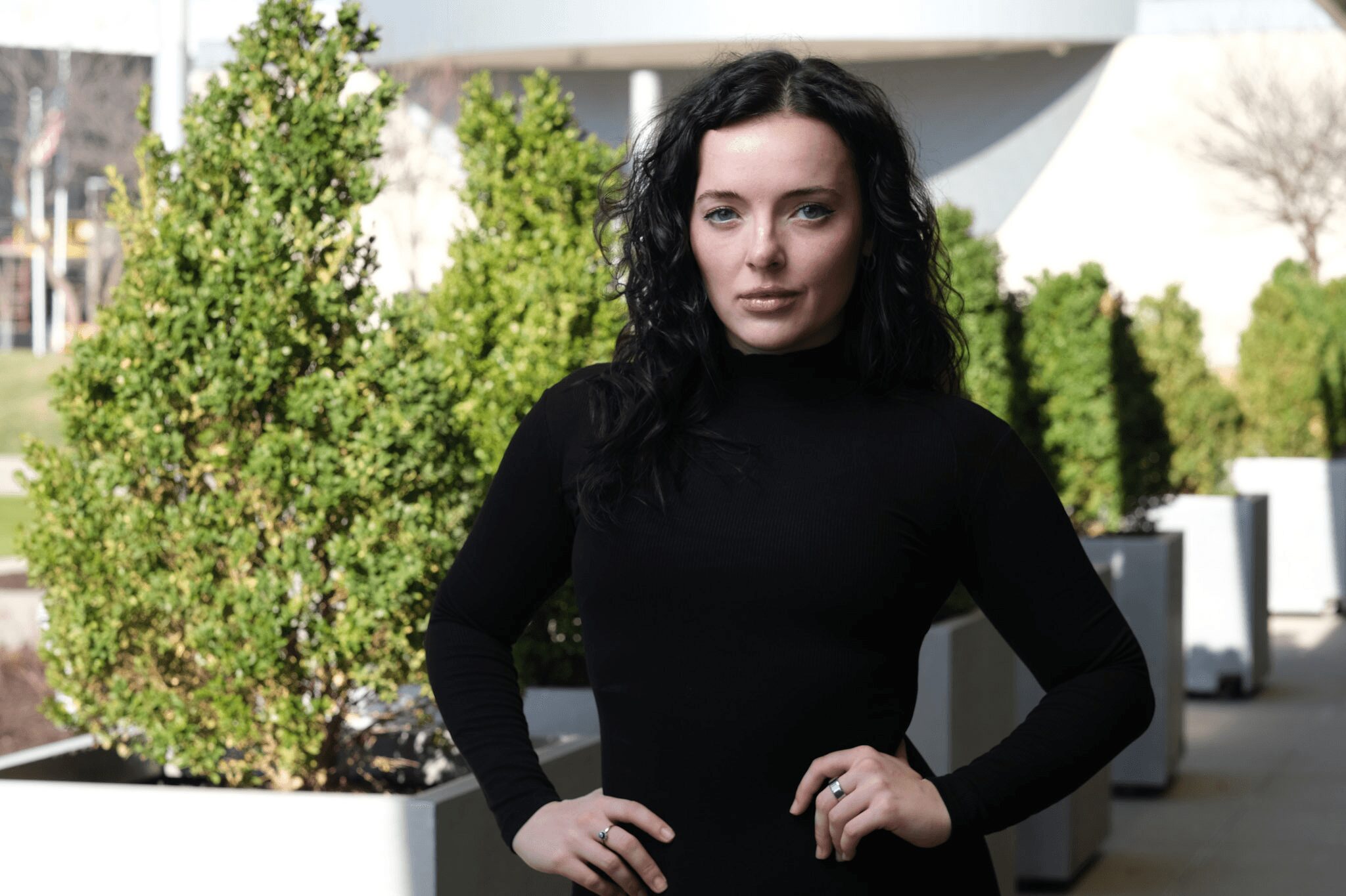 Woman with dark curly hair wearing a black turtleneck, standing outdoors with green bushes in background.