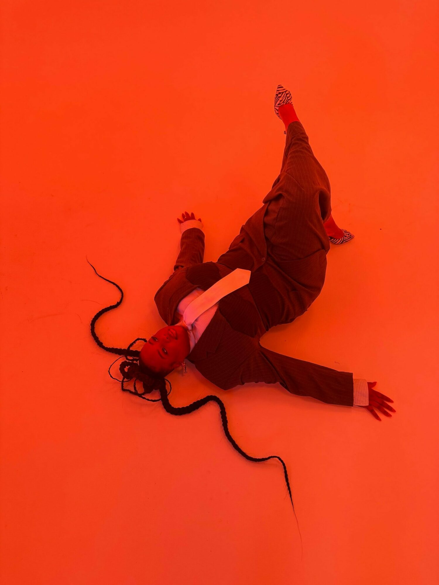 Person lying on the floor with a red background, head near a black cord, wearing dark clothing, in a relaxed pose.