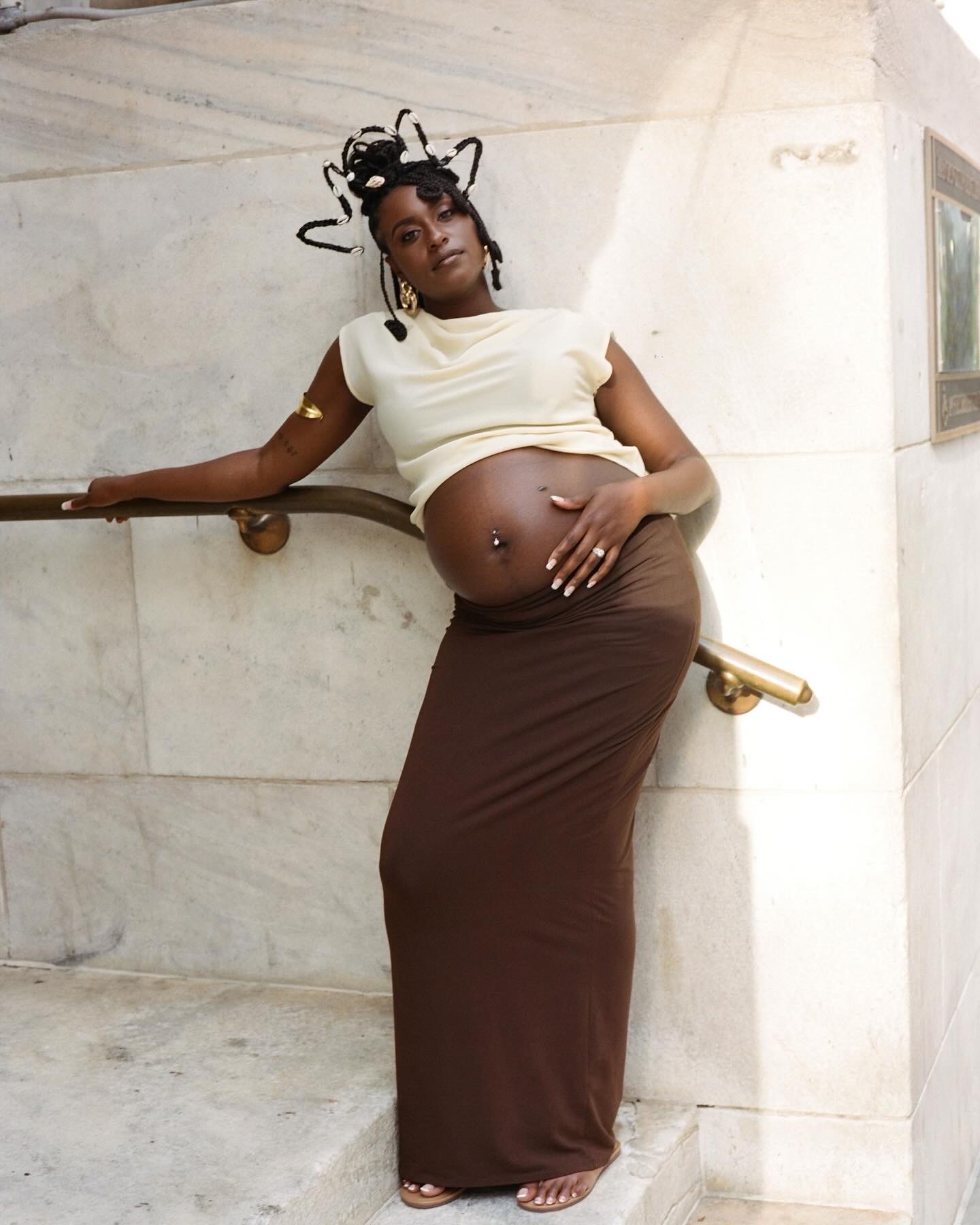 Woman with styled hair and a belly button piercing leans against a wall, wearing a white top and brown skirt.