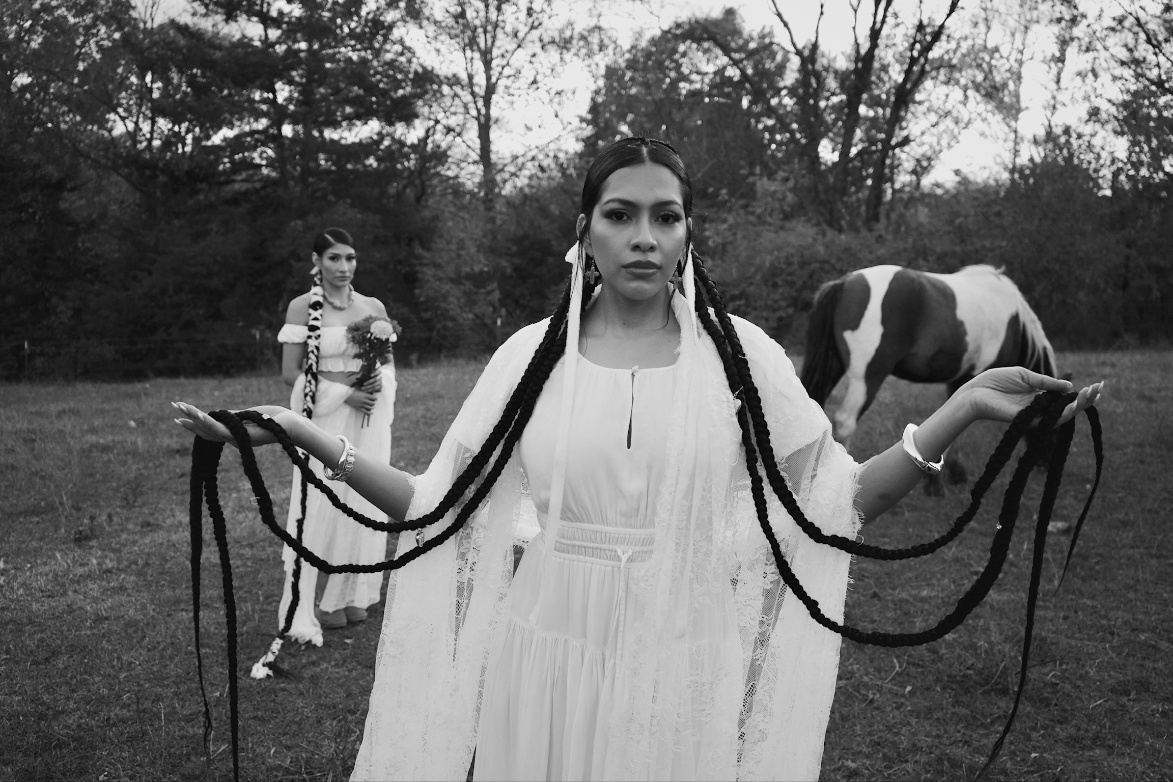 Woman with long braided hair in a white dress holding her braids outdoors, with a person and a cow in the background.