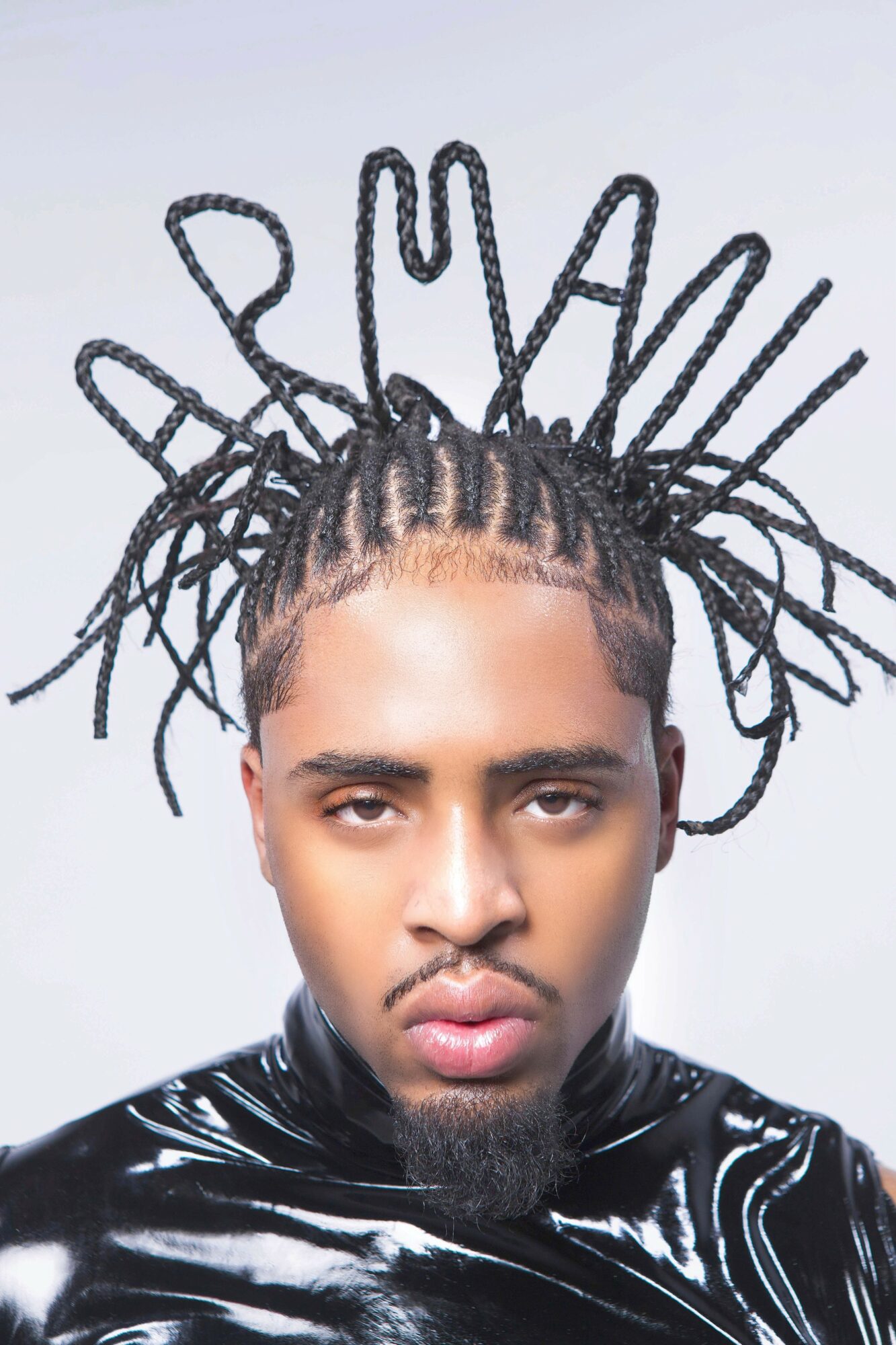 Person with styled dreadlocks arranged in a crown-like pattern, wearing a shiny black top, facing forward.