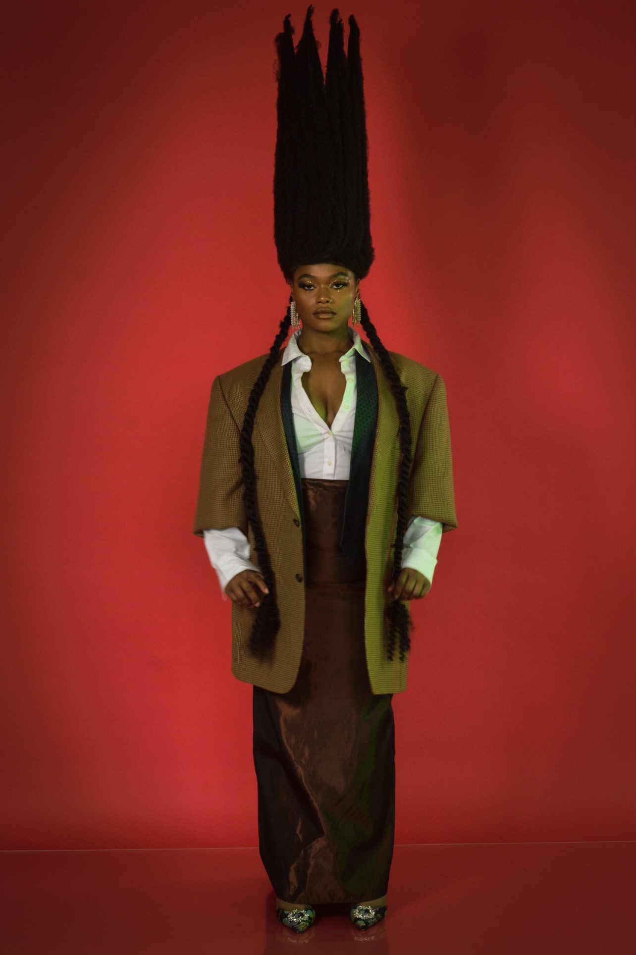 Person with tall, black hairstyle, wearing a brown coat over a white shirt, standing against a red background.