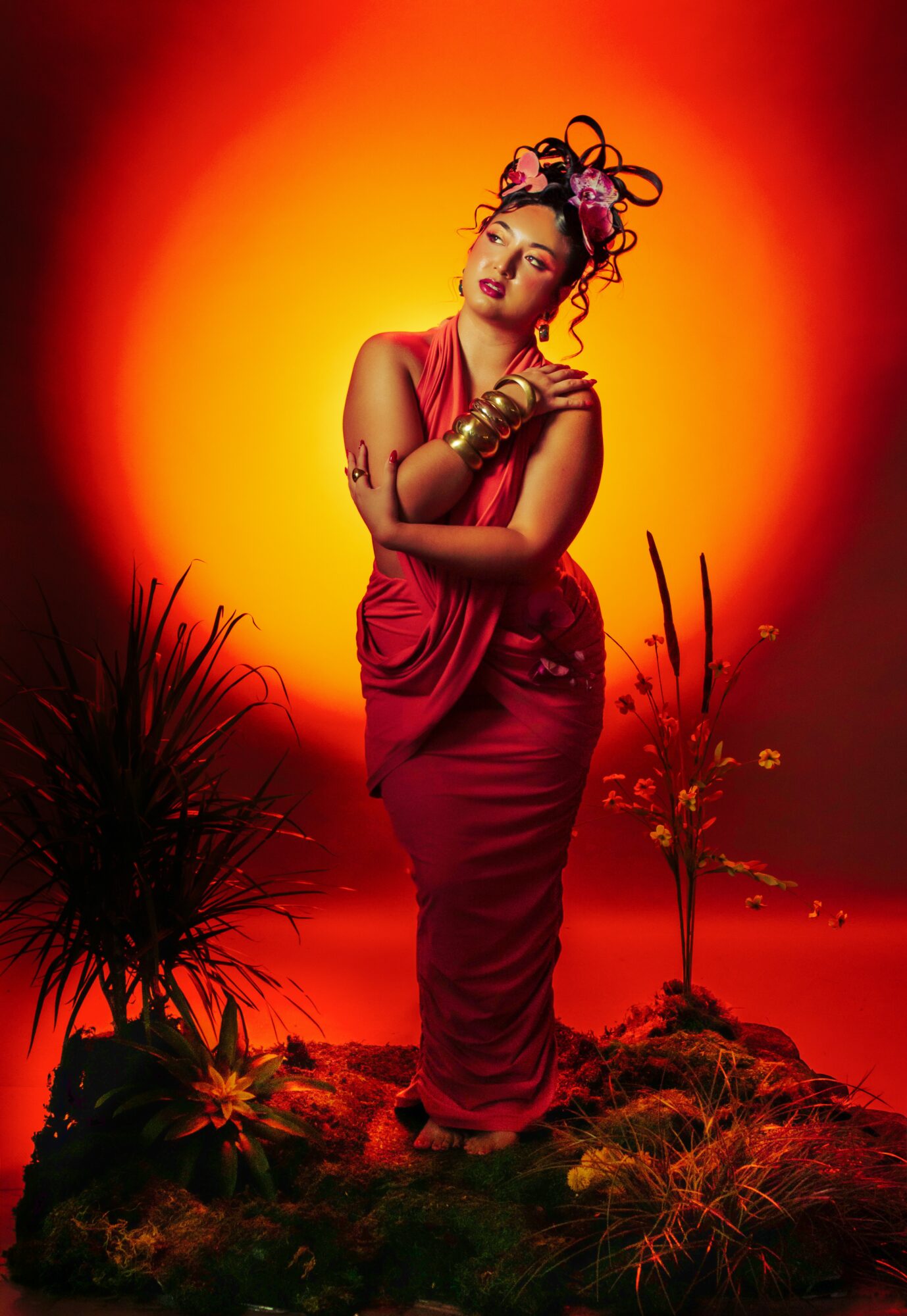 Woman in a red dress with elaborate hair and jewelry stands against a fiery orange background with plants around her.