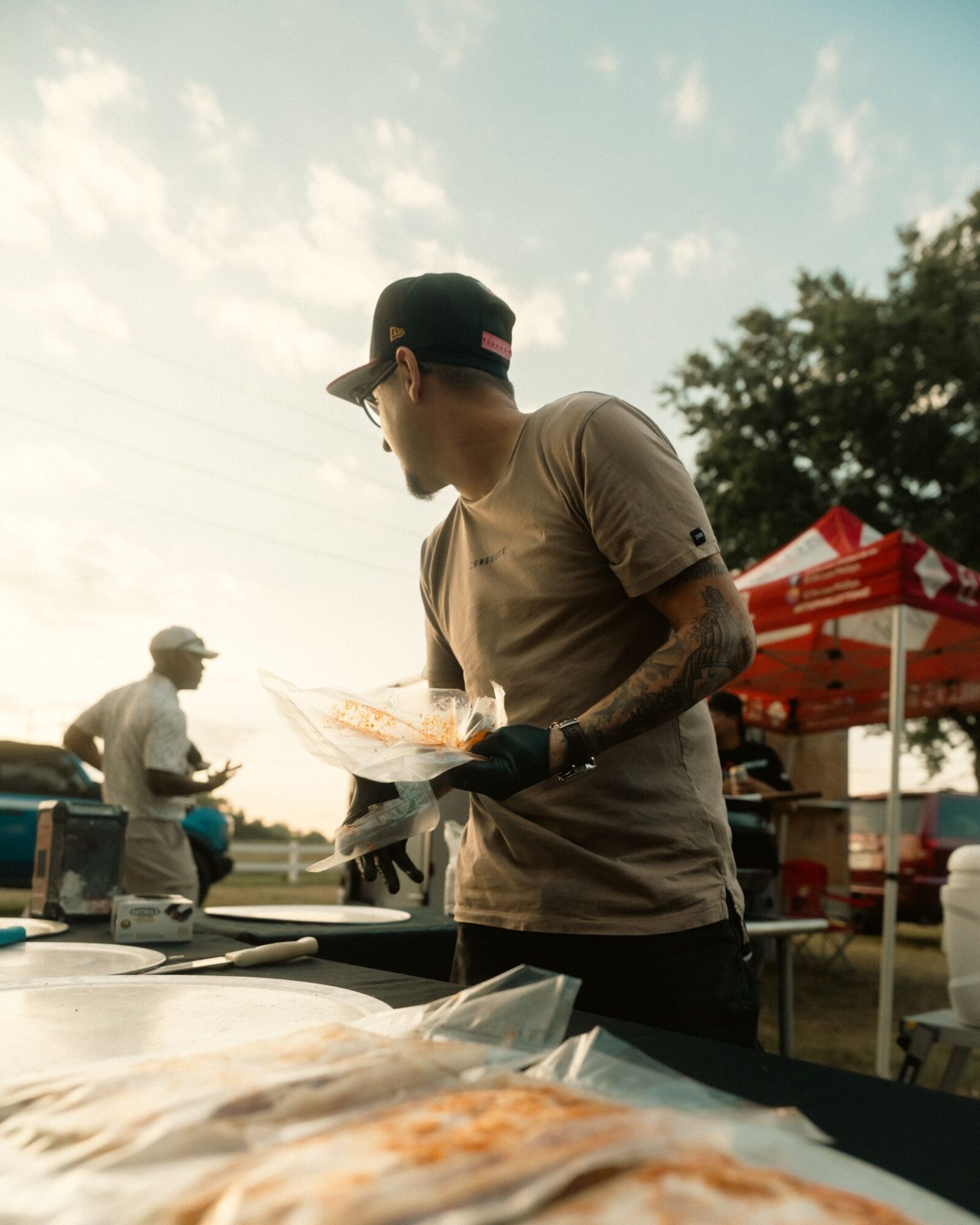 Person wearing a cap and sunglasses preparing food at an outdoor event, with tents and other people in the background.