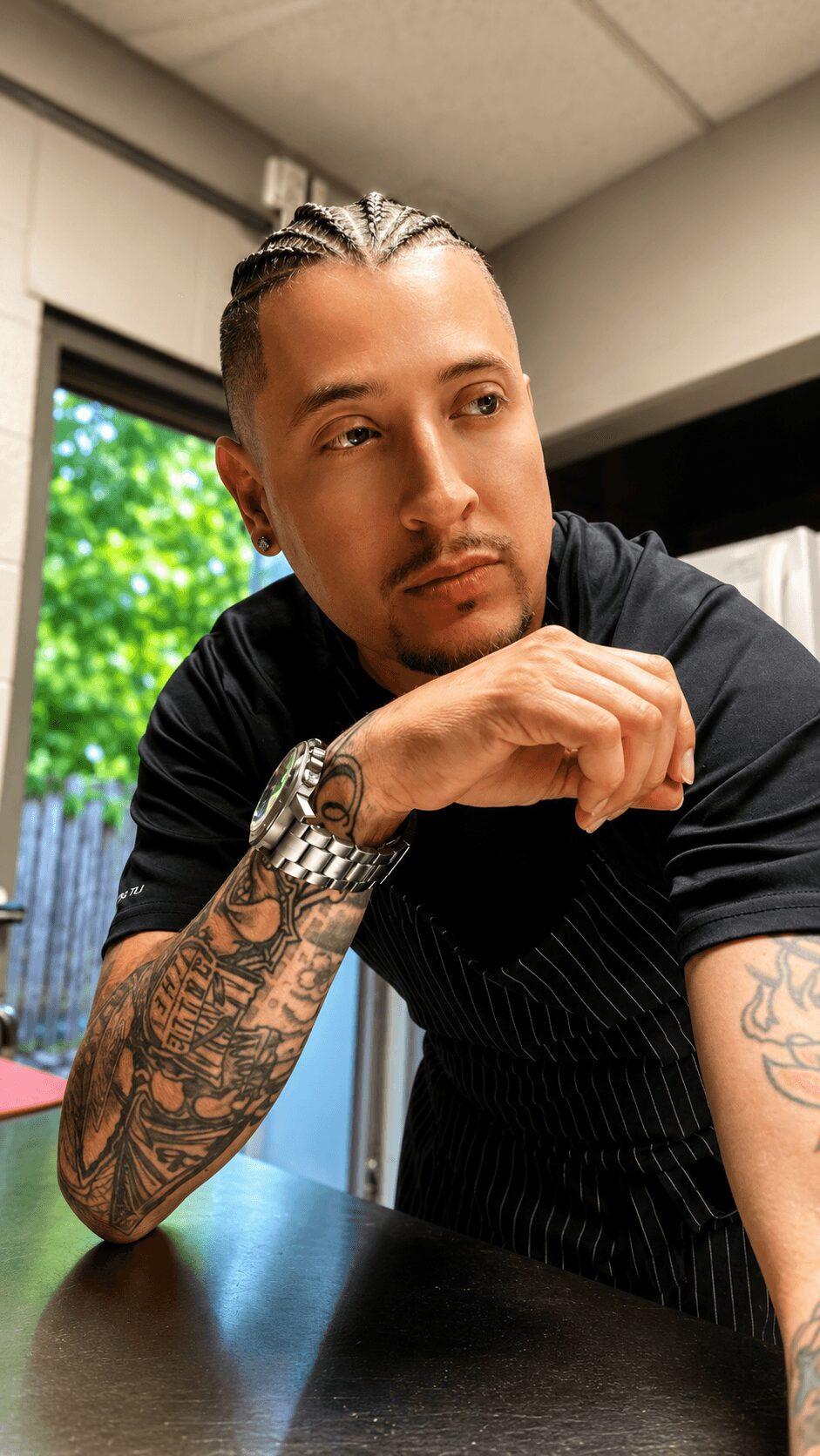 Young man with slicked-back hair and tattoos, resting his chin on his hand, indoors with a window showing greenery outside