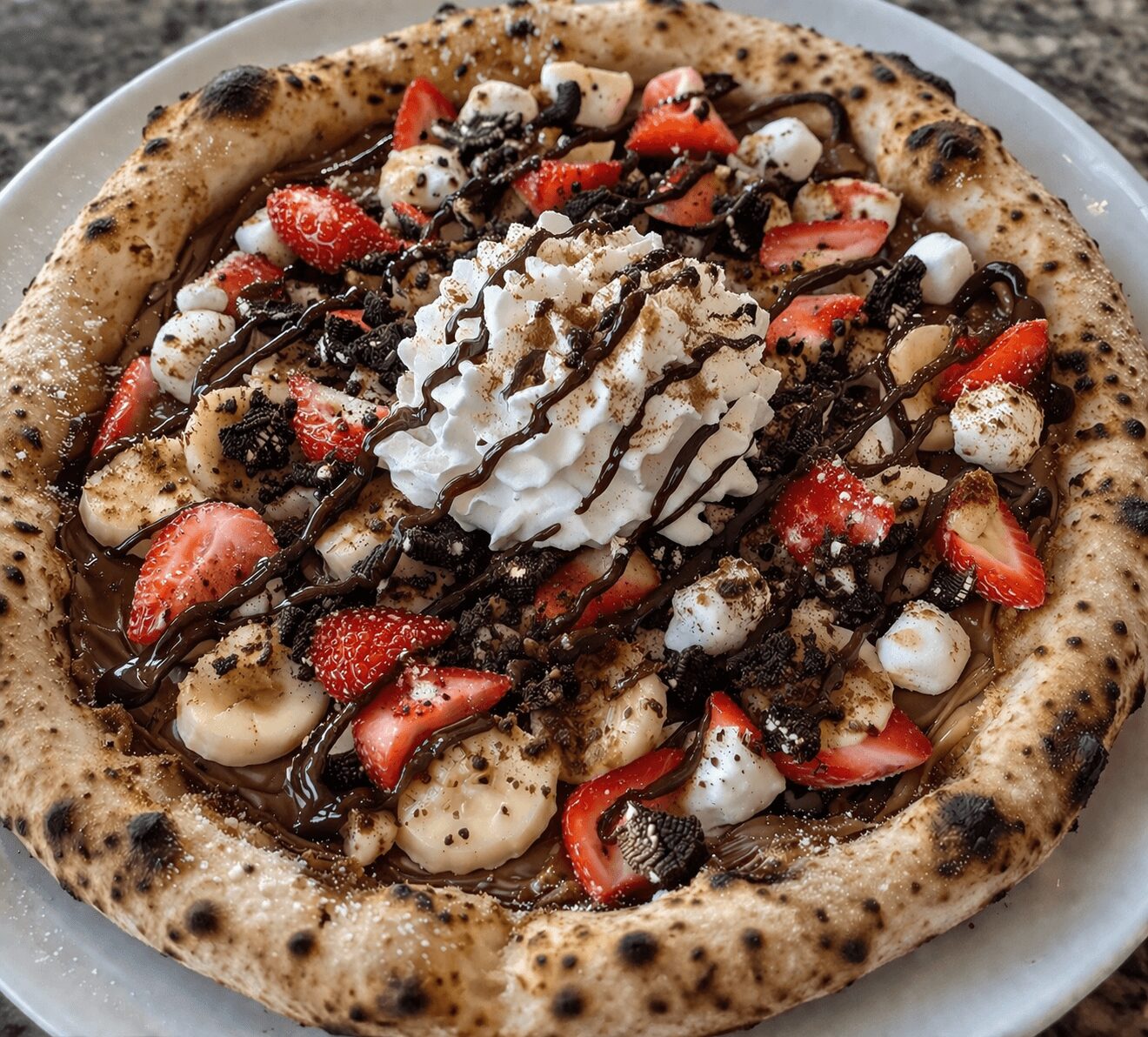 Pizza topped with strawberries, bananas, chocolate drizzle, whipped cream, and cookie crumbs on a plate.
