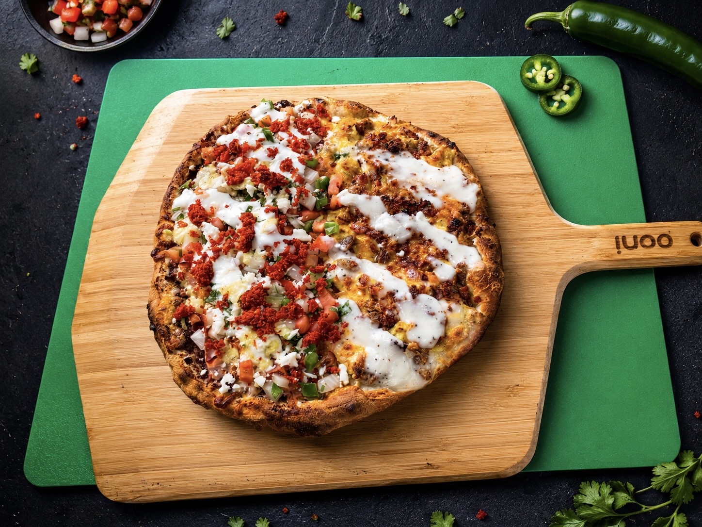 Half pizza on a wooden board with toppings and sauce, on a green cutting mat, with jalapenos and herbs nearby.