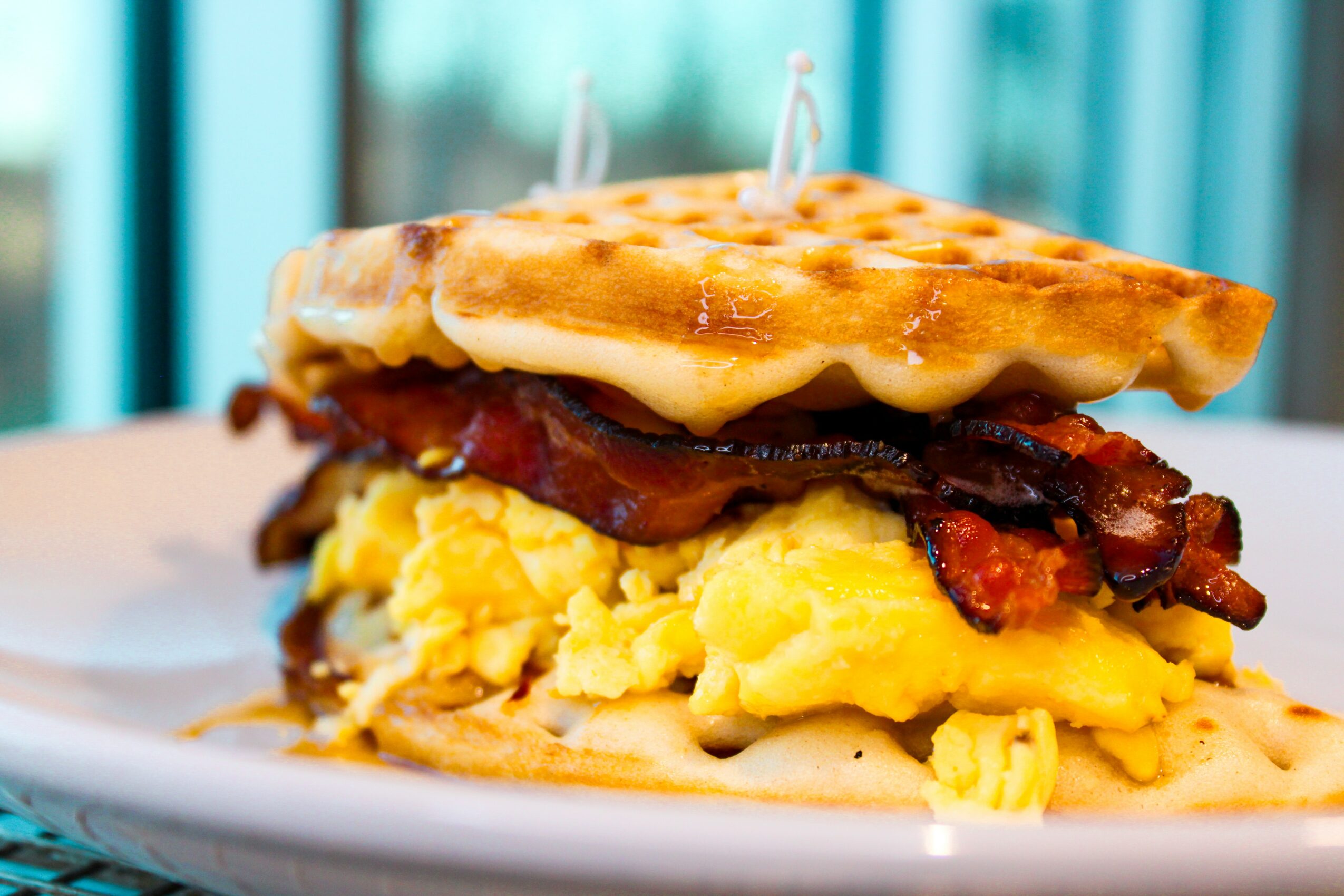 A breakfast sandwich with scrambled eggs, bacon, and cheese on toasted bread, on a white plate, with a blurred background.