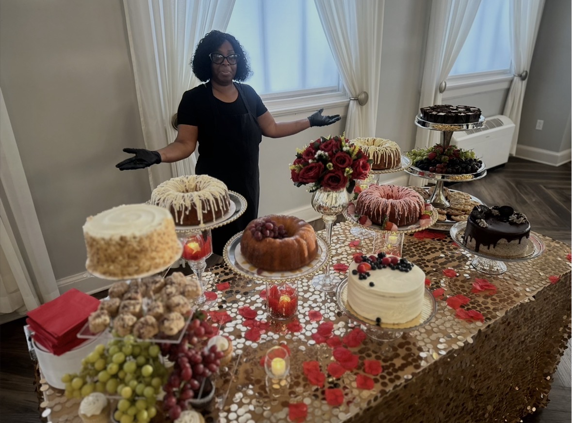 Person standing behind a table with various cakes and desserts, in a room with large windows and curtains.
