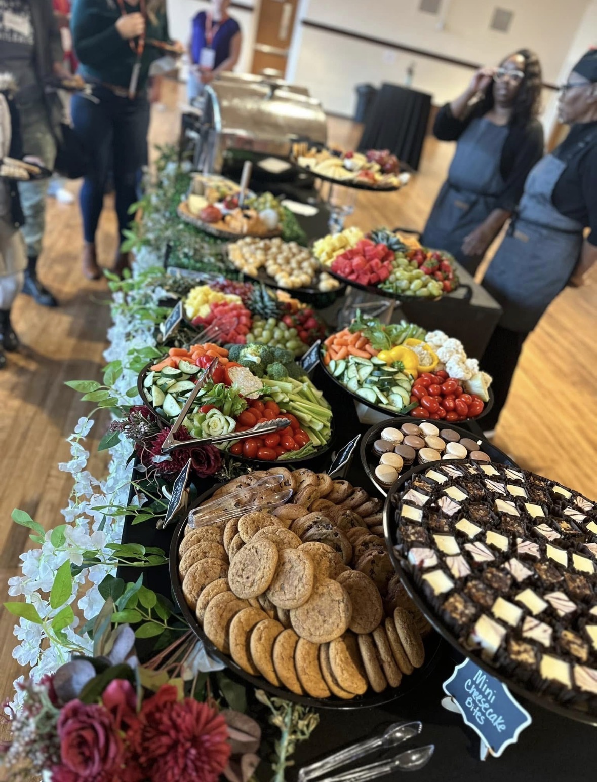 Table with assorted desserts and fruit, people in background, indoor setting, greenery decoration, some people taking photos.