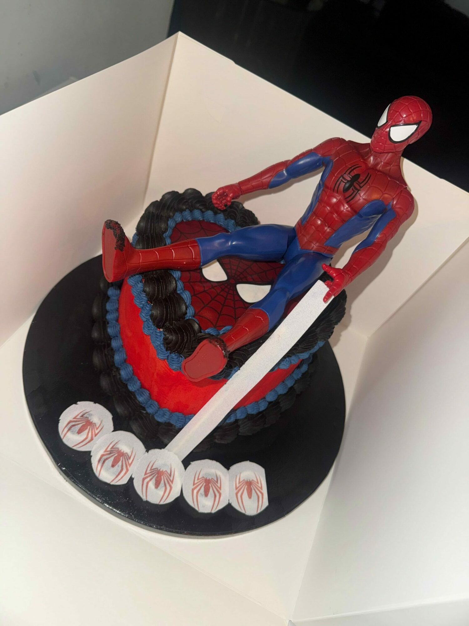 Spider-Man figurine on a round black cake with red and white decorations, holding a white sword-like object.