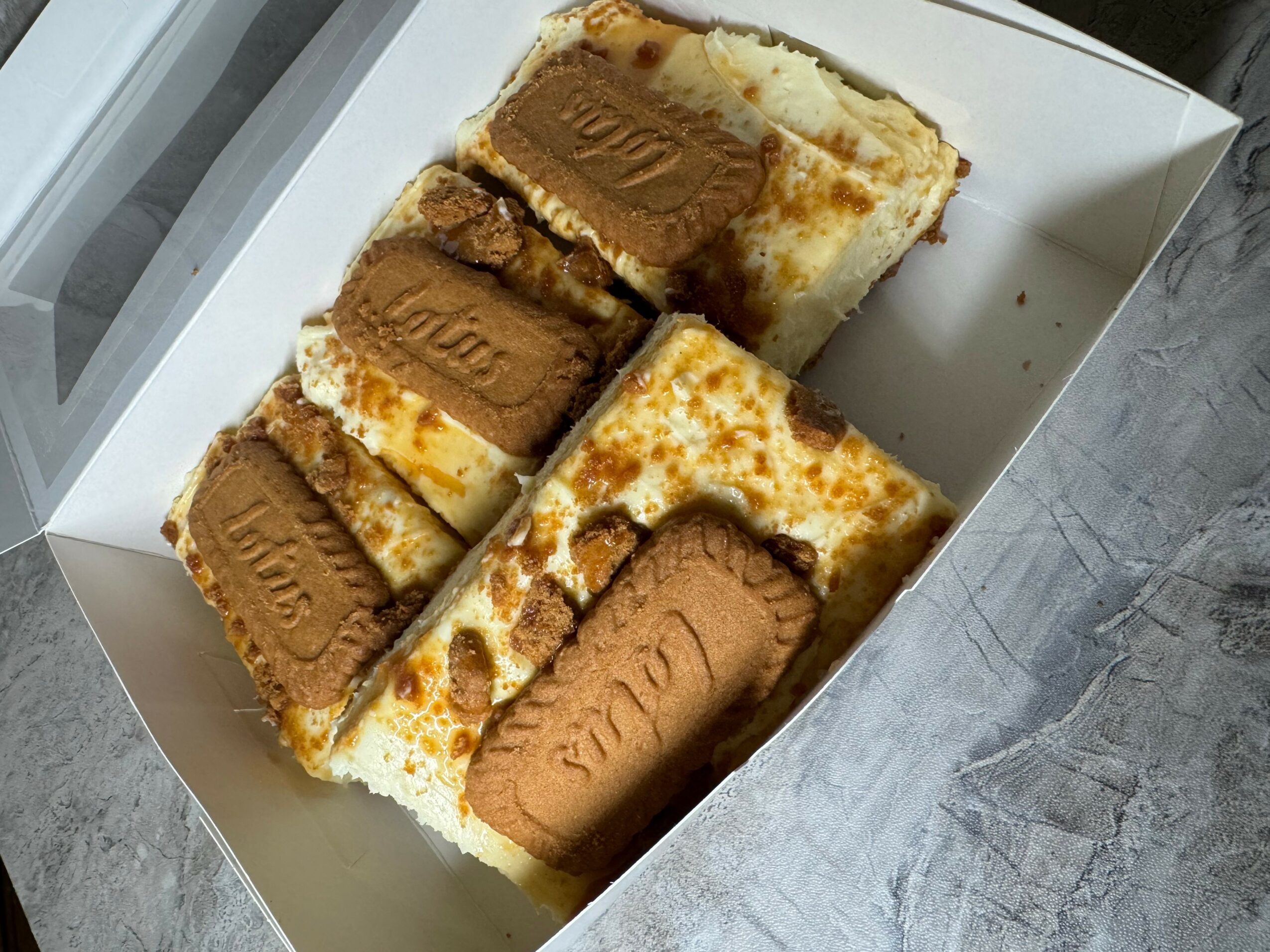 Four slices of layered dessert with cream, chocolate, and caramel, in a white box on a gray surface.
