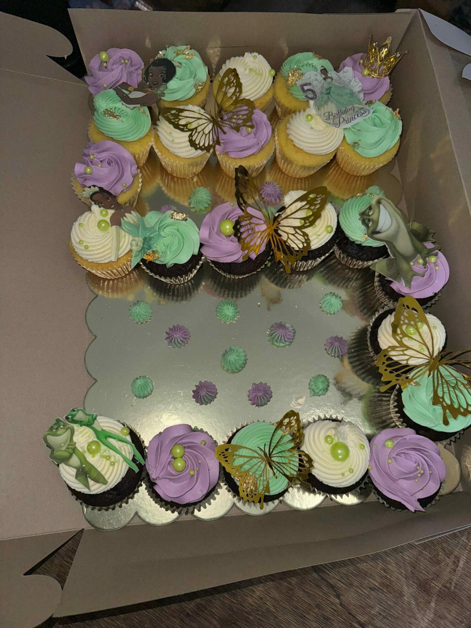 Box of cupcakes decorated with pastel-colored frosting and butterfly toppers, arranged in a circular pattern inside the box.