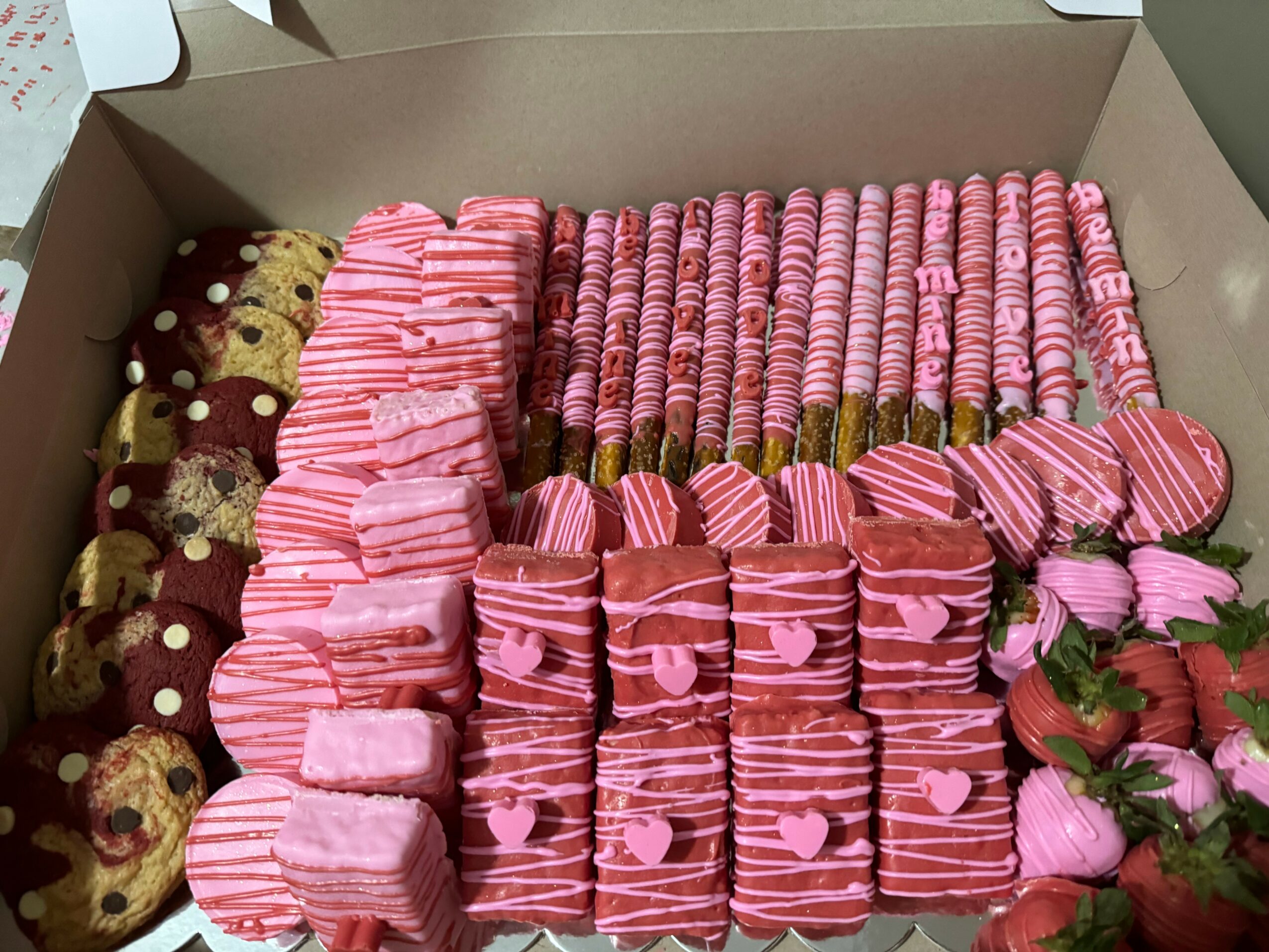 Box filled with pink and brown decorated cookies, some with hearts and stripes, and a few with strawberries, arranged in rows.