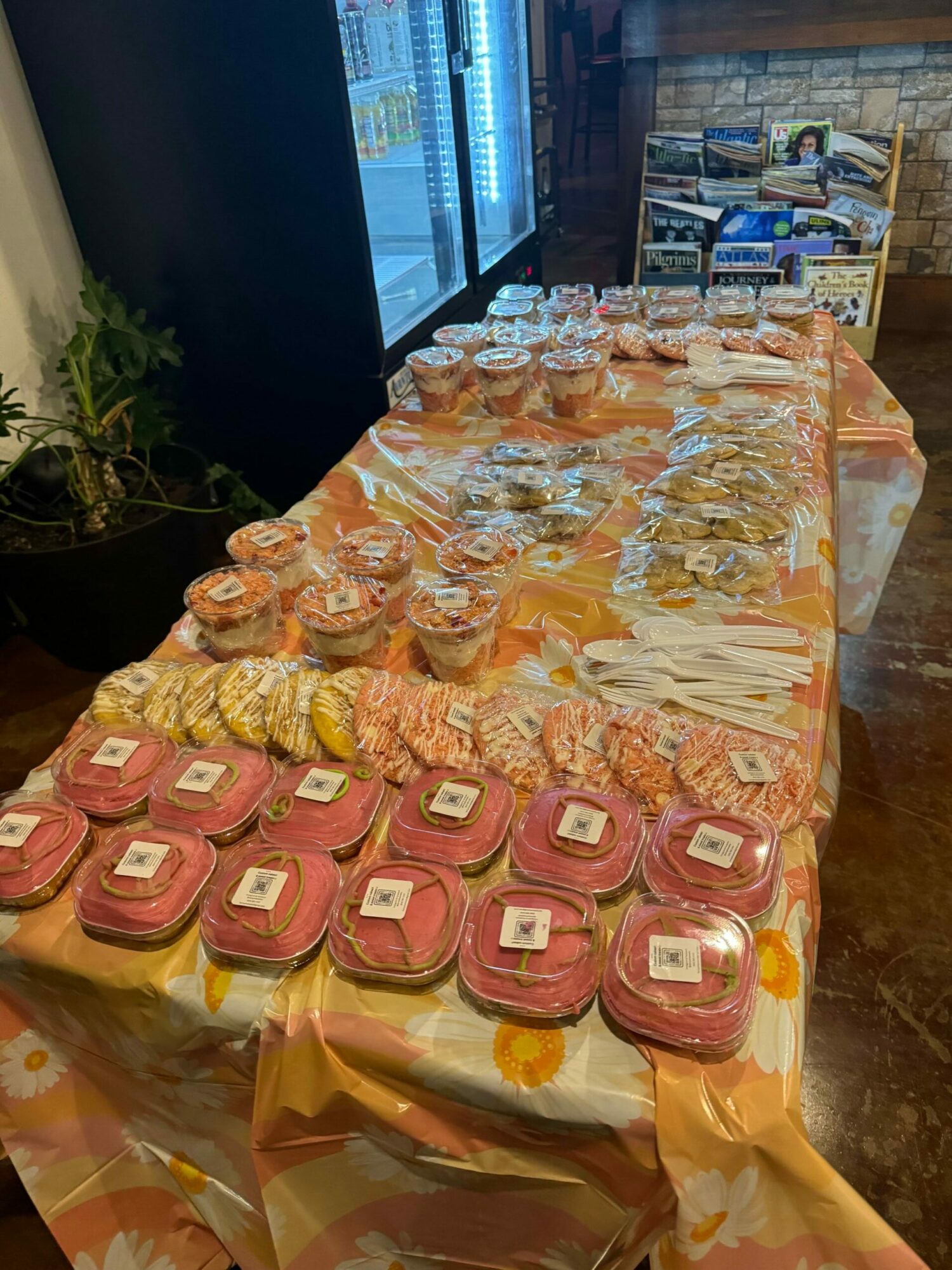 Table with pink and yellow wrapped cakes and snacks, decorated with orange and yellow paper, in a room with a window and shelves.