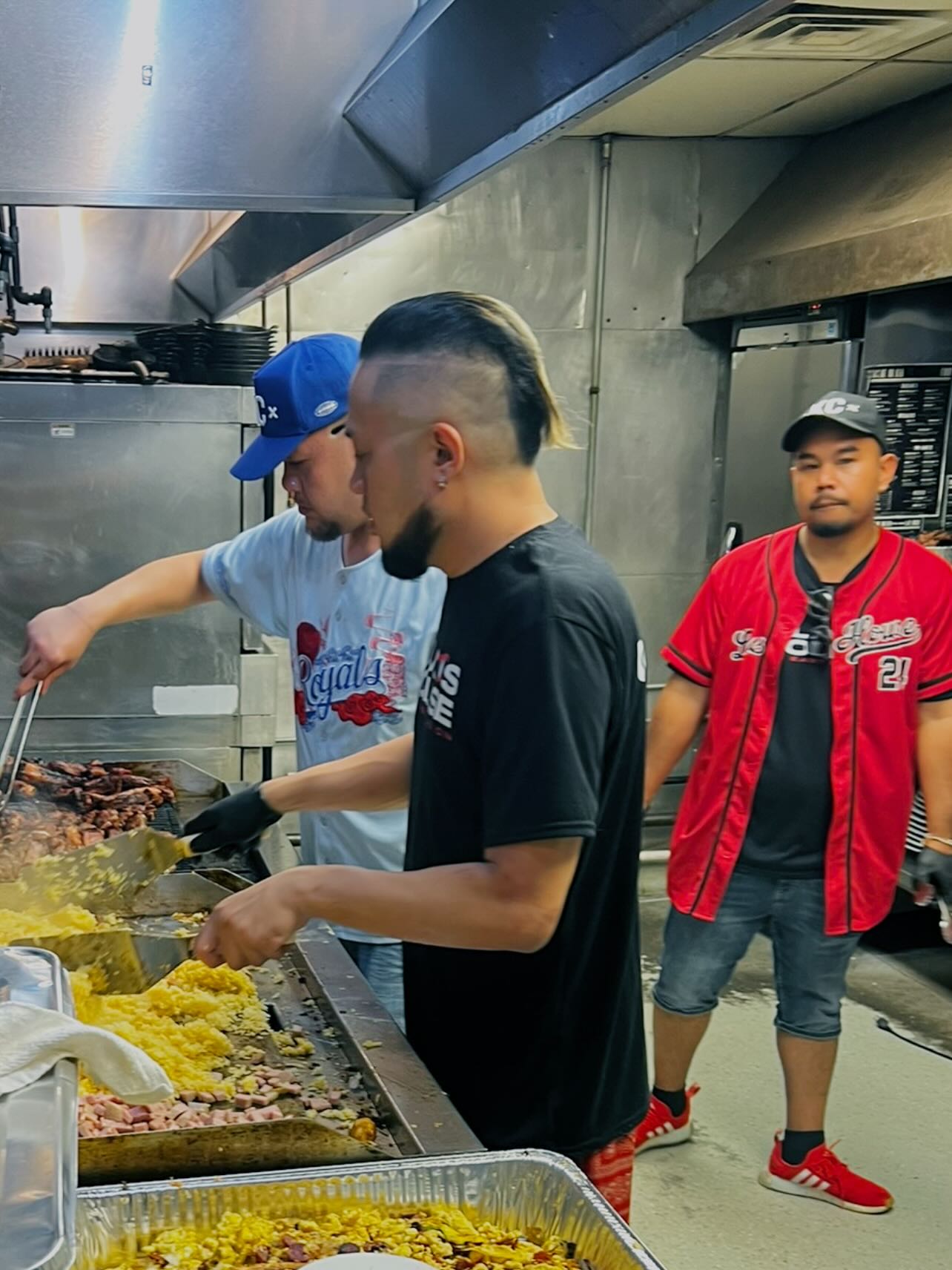 Three young men preparing food in a commercial kitchen, one wearing a black shirt, another in a blue cap, and the third in a red jacket, with cooking equipment around them.