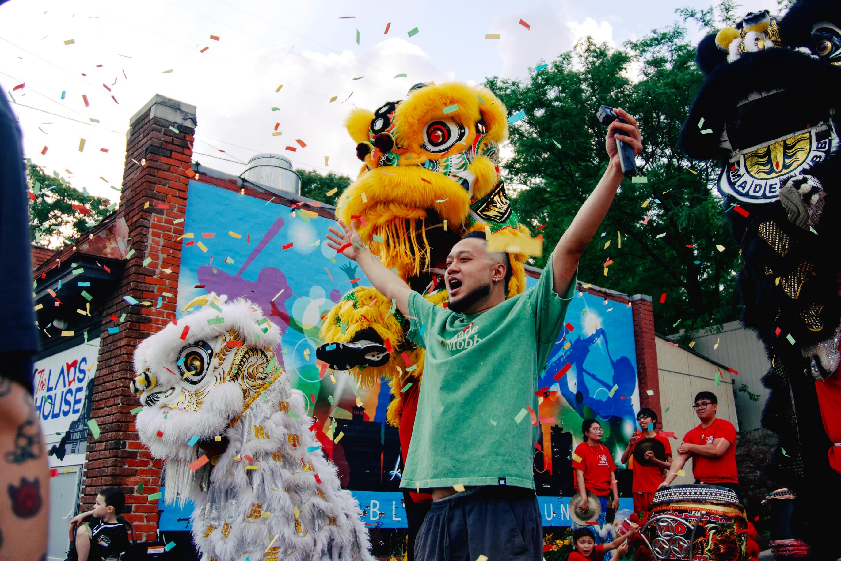 Person celebrating during a colorful parade with lion dance costumes and festive decorations, outdoors with trees and buildings in background.