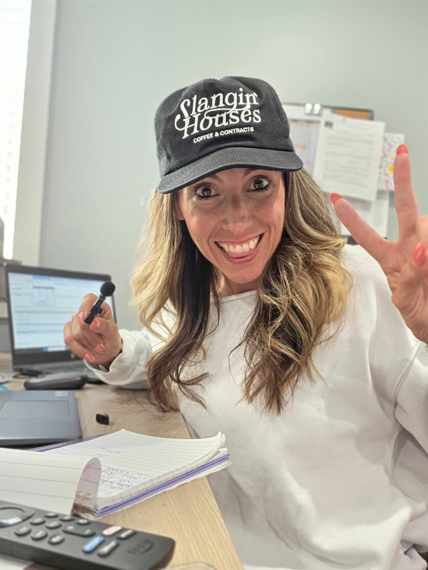 Woman wearing a cap, smiling, making a peace sign, sitting at a desk with a computer and notebooks.