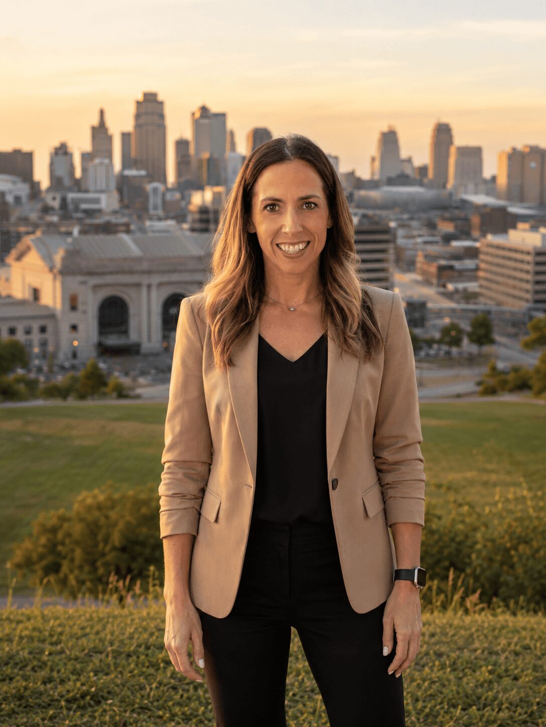 Woman standing outdoors with city skyline in the background during sunset, smiling, wearing a beige blazer and black top.