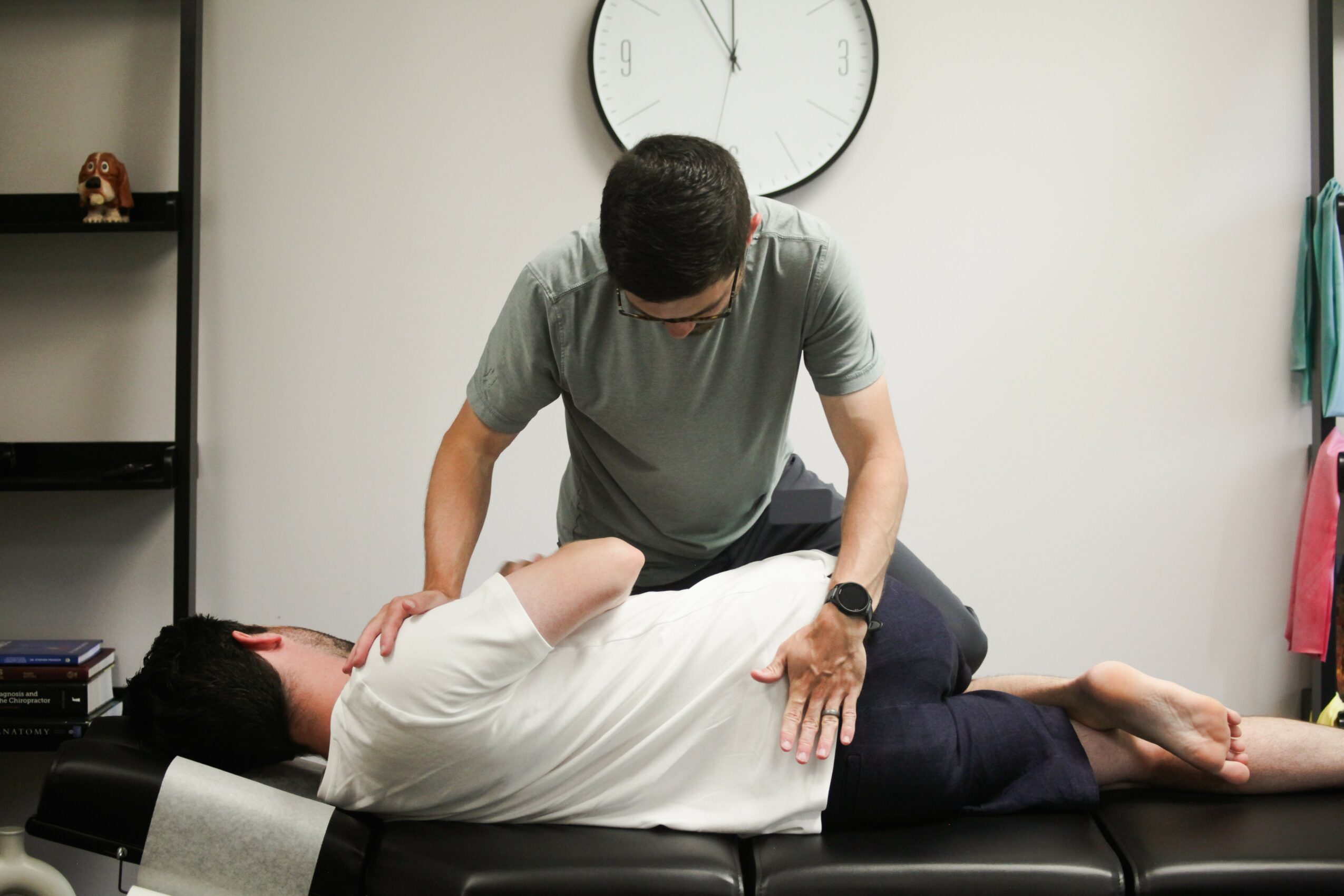 A person receiving a massage from a therapist in a room with a clock on the wall, shelves, and towels.