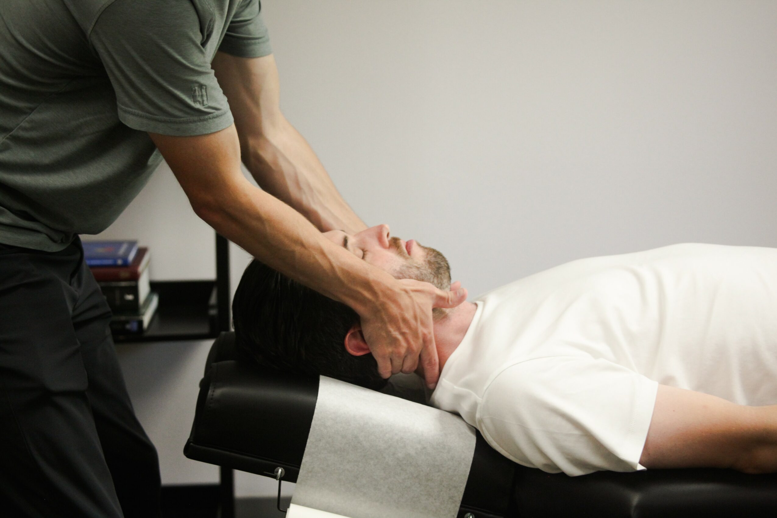 Person receiving neck massage from another person in a clinical setting, lying on a table with a white shirt.