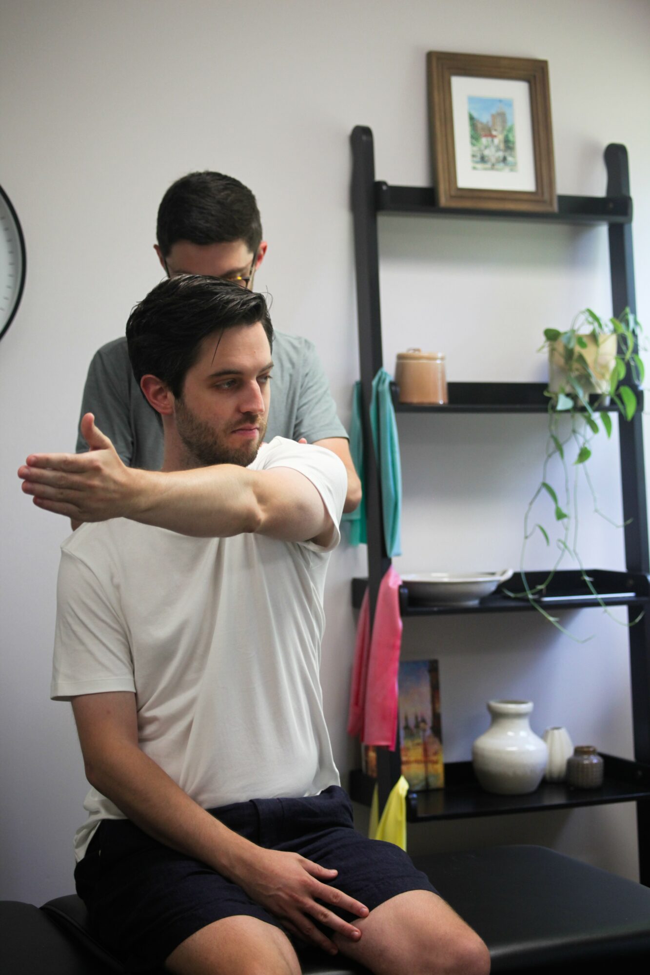 Two men in a room, one sitting and stretching arm, the other standing behind. Shelves with decor and a framed picture on wall.