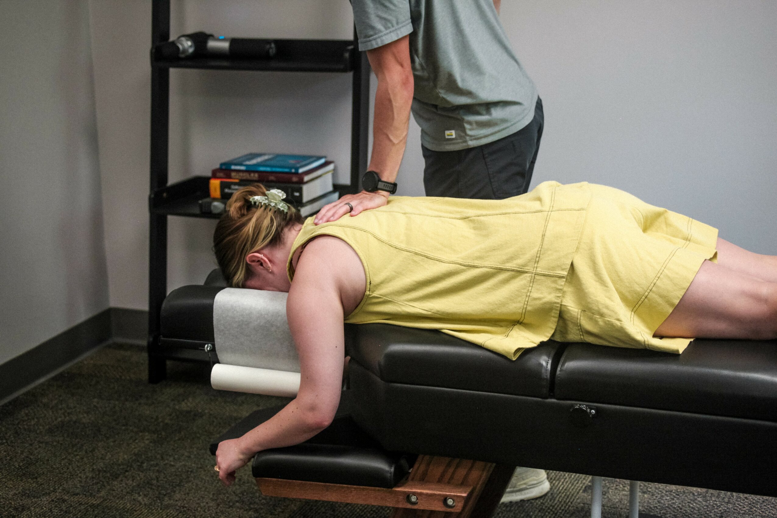 Person lying face down on a massage table with a yellow dress, in a room with a black shelf and gray walls.