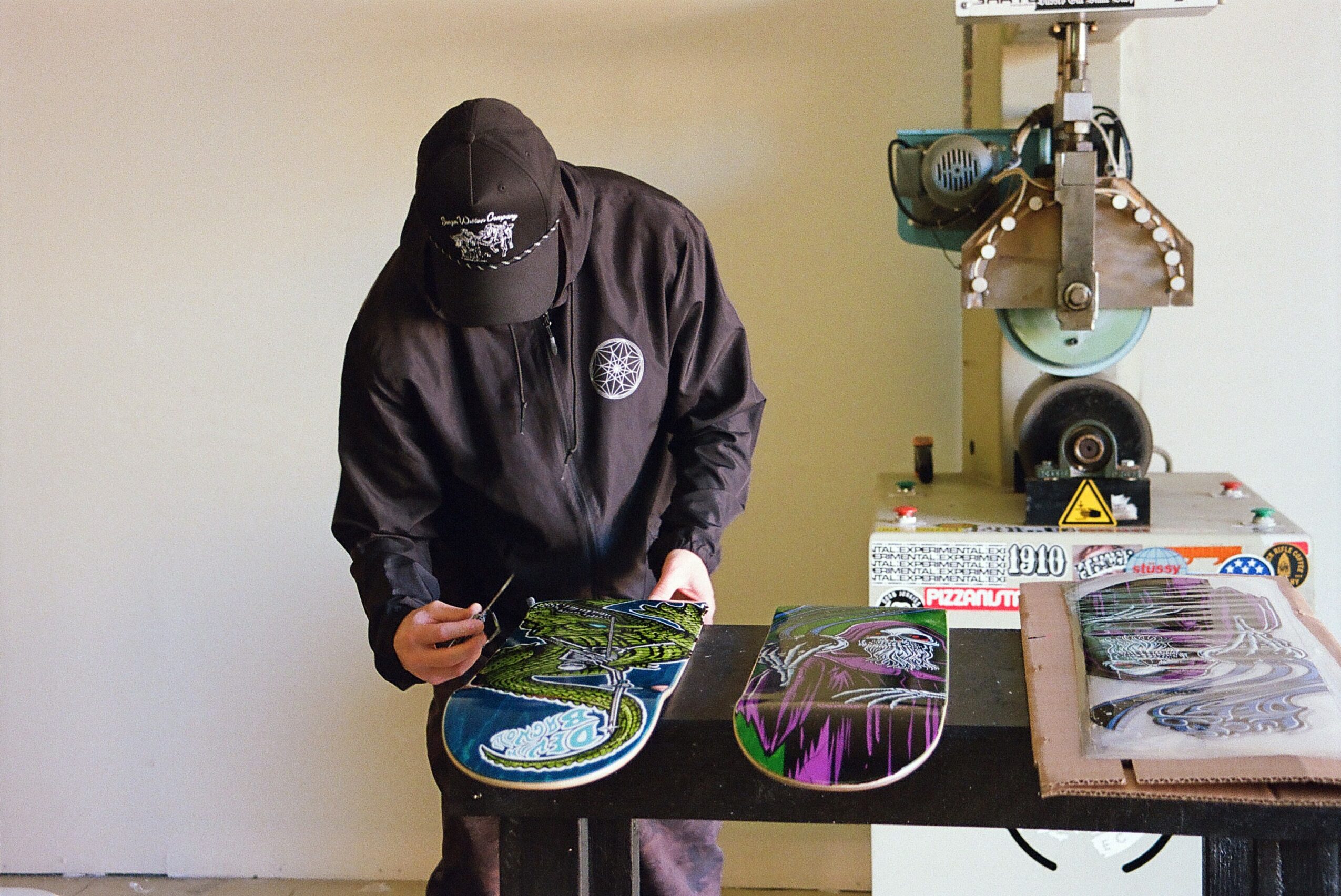 Person wearing a black jacket and cap working on skateboard decks at a table, with equipment in the background.