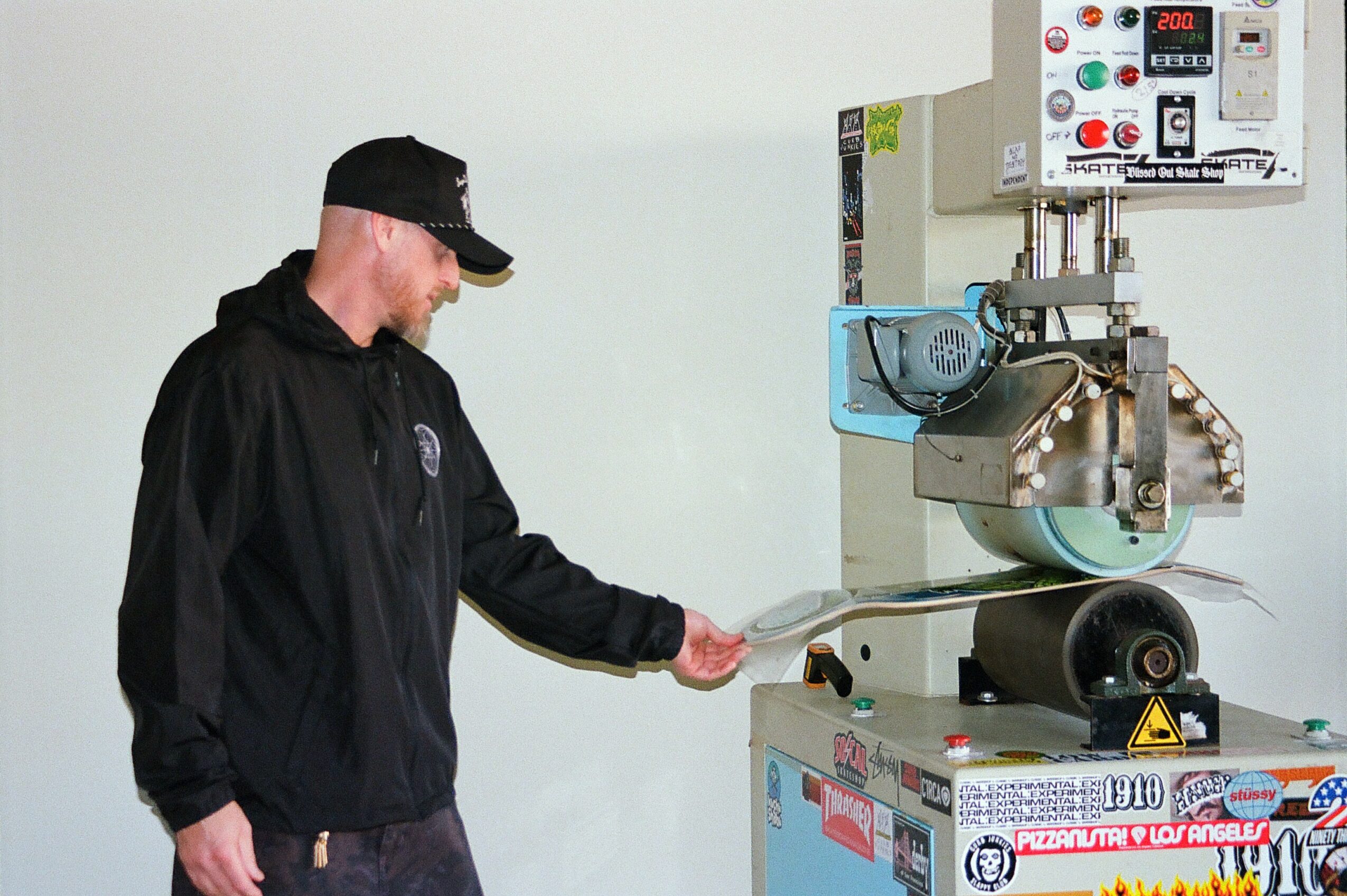 Man operating a large industrial machine in a room with a plain light-colored wall, wearing a black jacket and cap.