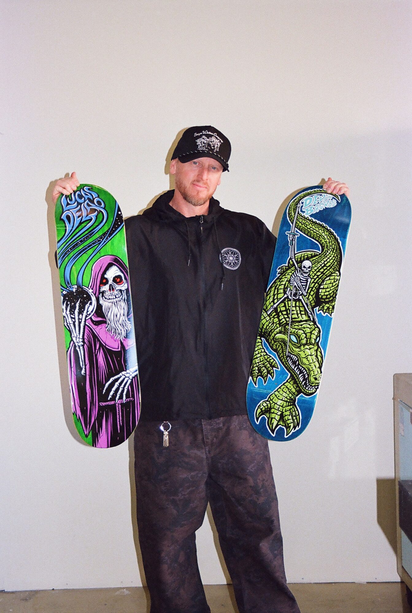 Person holding two skateboards, one with a colorful skeleton design and the other with a green and blue crocodile pattern, standing against a plain wall.