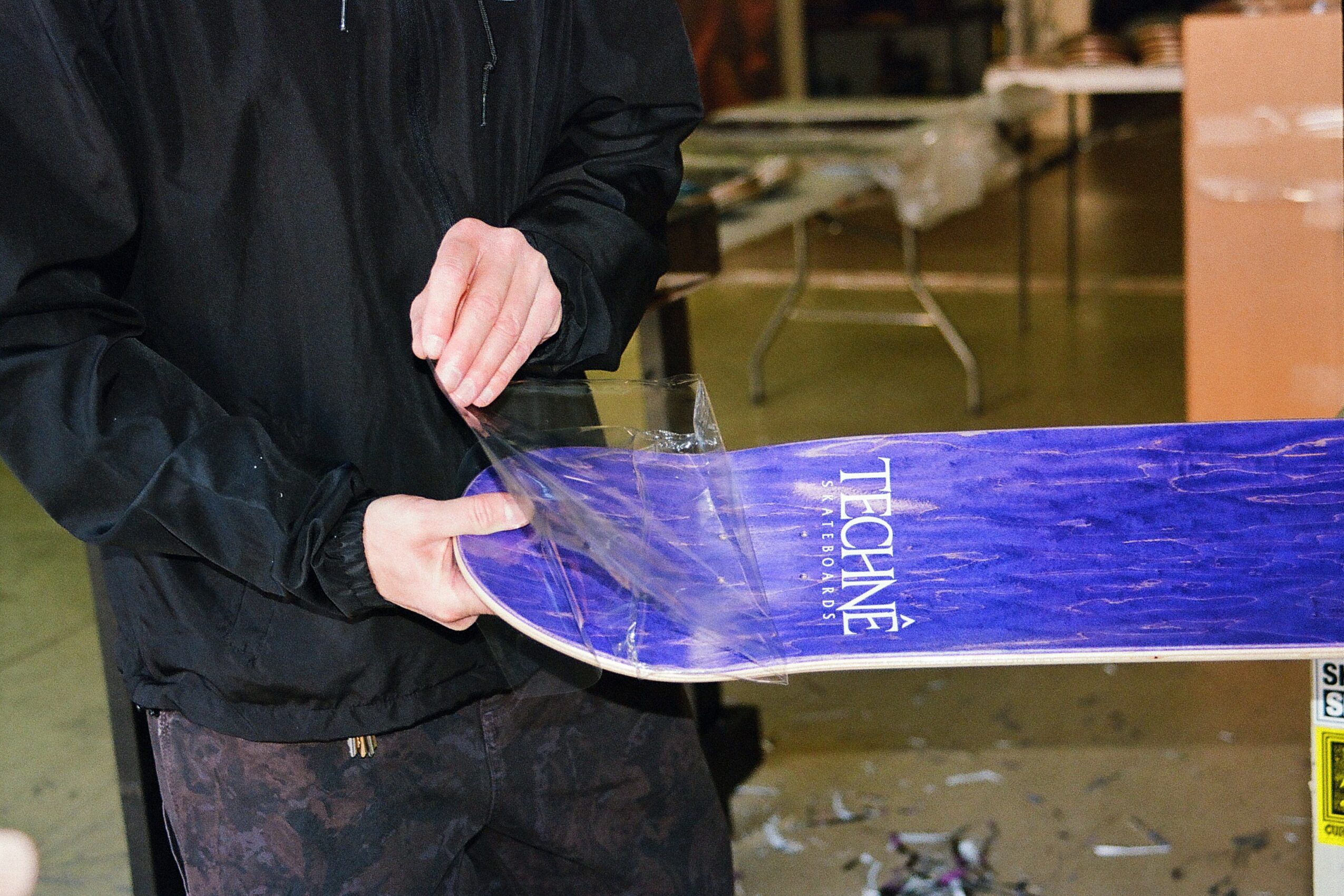 Person holding a blue snowboard with white text, in an indoor setting with boxes and equipment in the background.