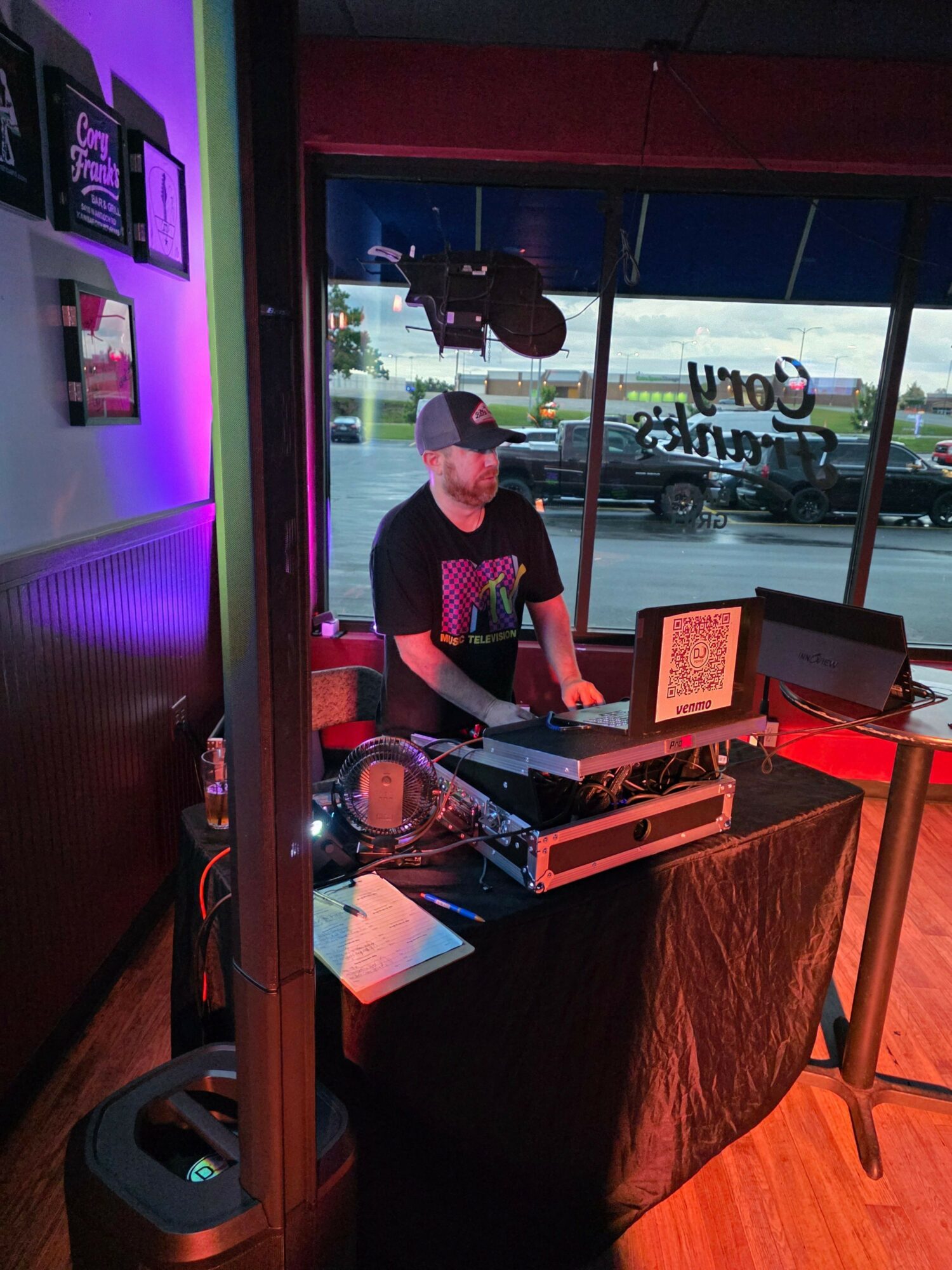 Person wearing a mask working with DJ equipment in a room with purple lighting, window showing outdoor scene.