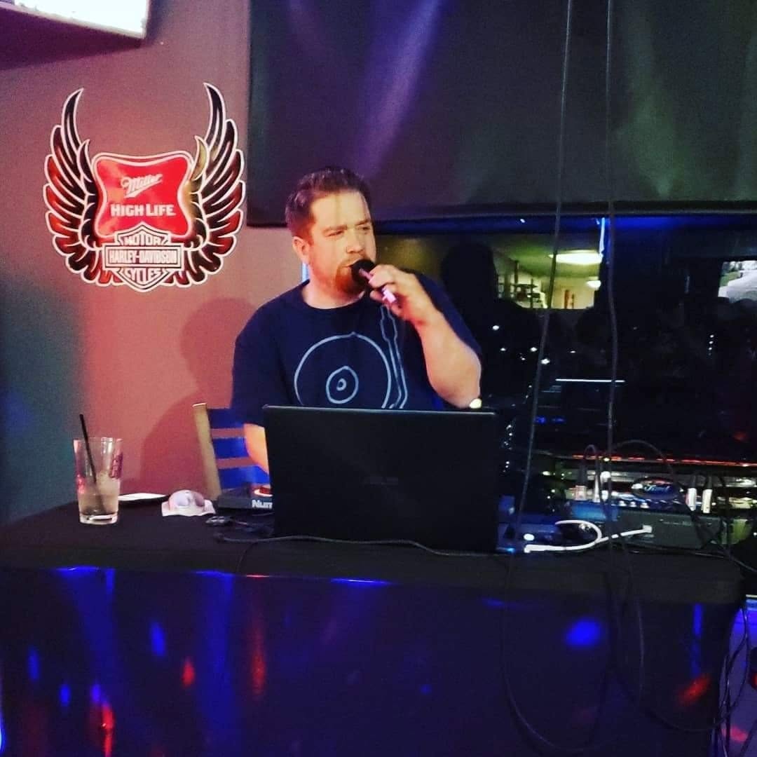 Person singing into a microphone at a DJ booth with a laptop, in a dimly lit venue with a logo on the wall.