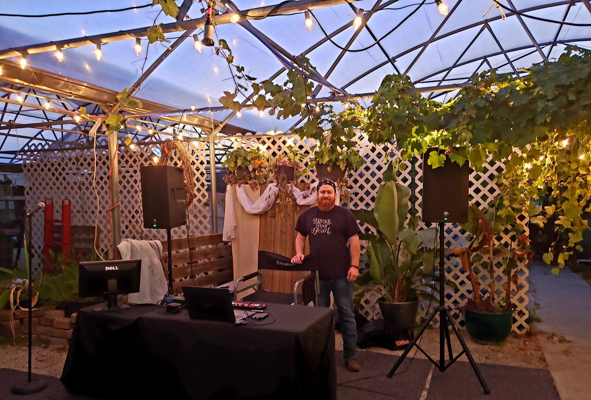 People standing under a decorated outdoor canopy with string lights, plants, and a table with equipment.