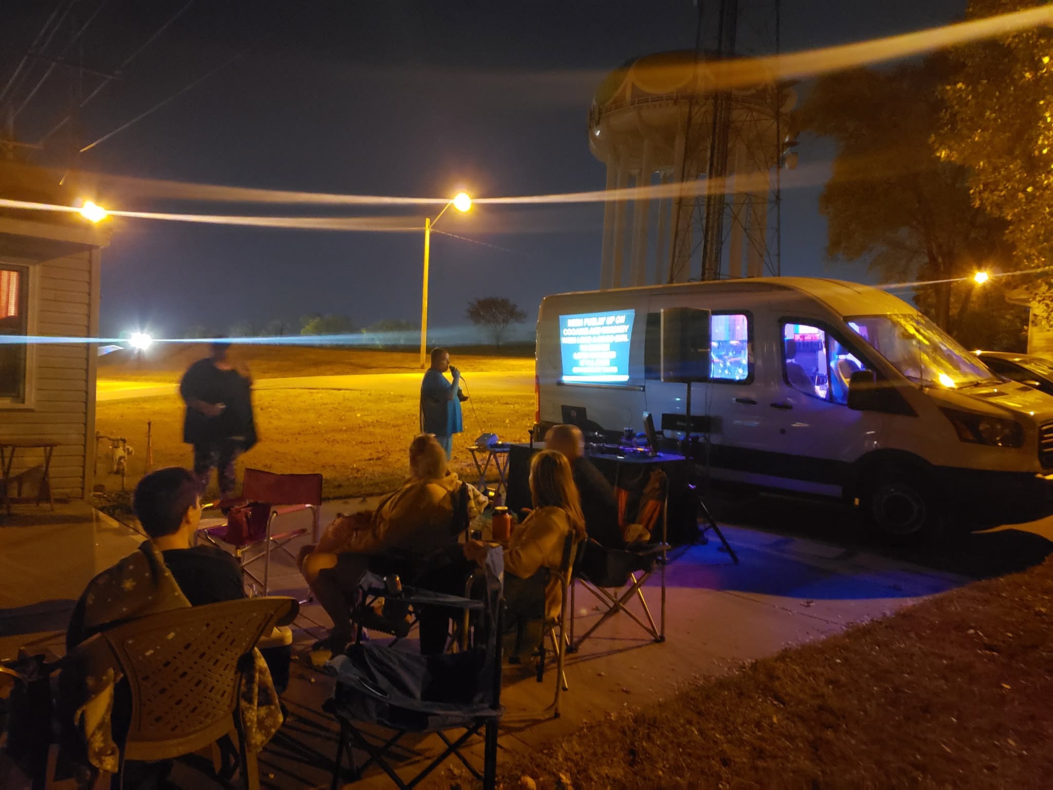 People sitting outdoors near a van with screens, nighttime scene with streetlights and a building in the background.