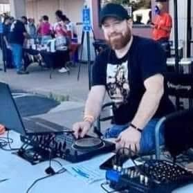 Man with a beard wearing a black cap and T-shirt, operating DJ equipment outdoors, with people in the background.