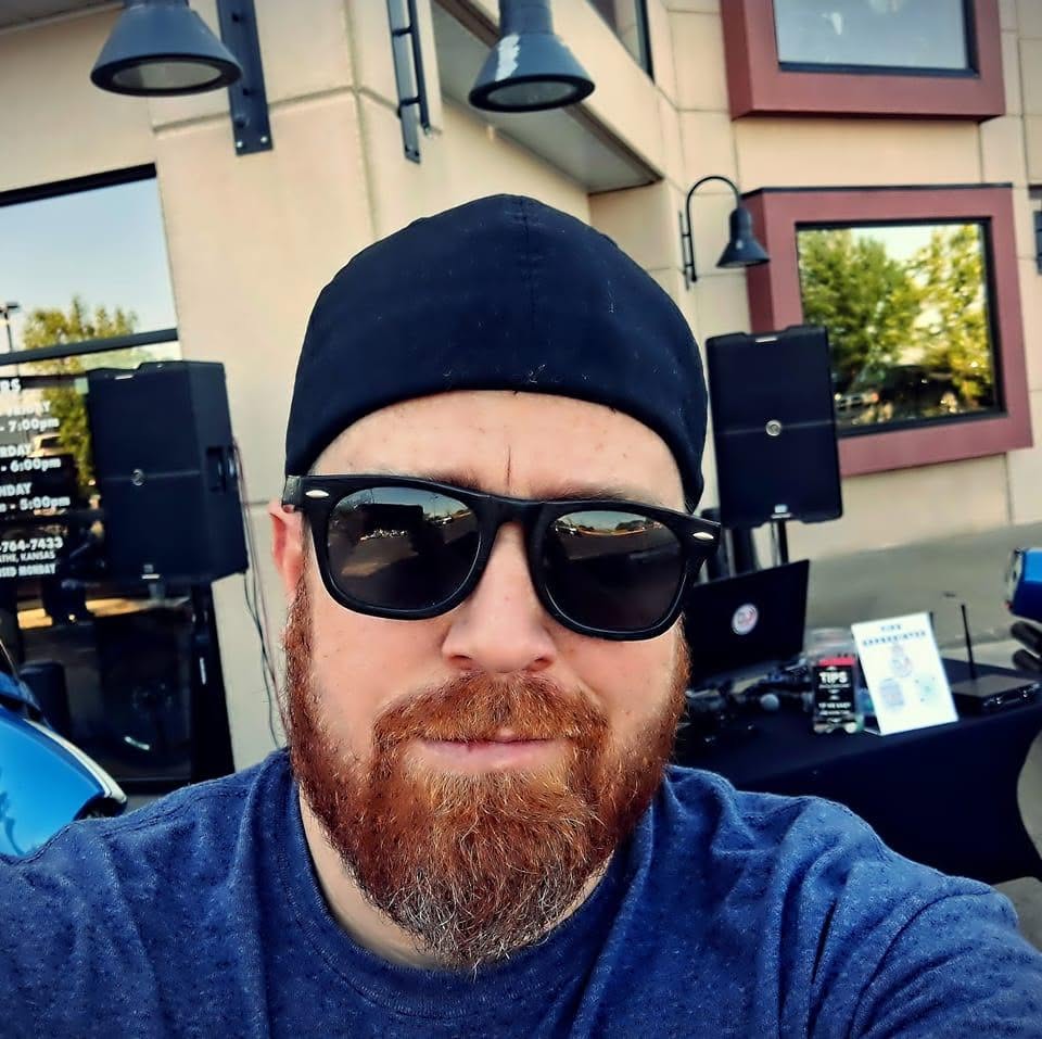Man with beard wearing sunglasses and a black cap outdoors, with a building and trees in the background.