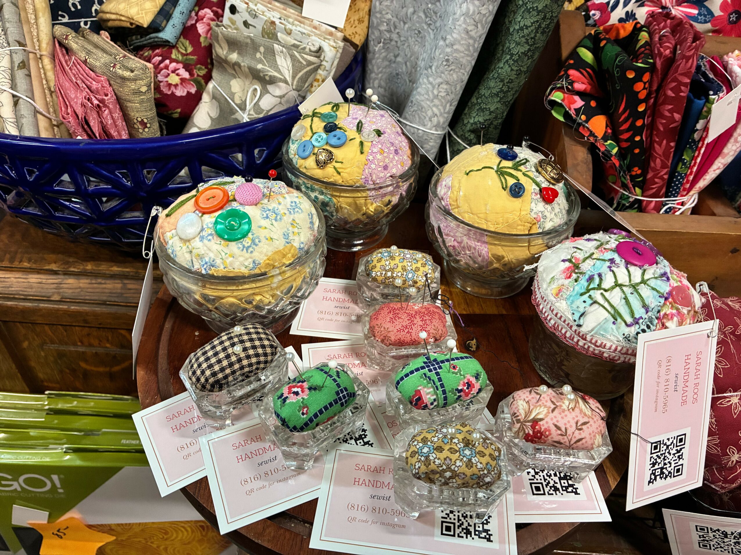 Display of colorful fabric items and small decorated fabric pouches on a table, with a blue basket in the background.