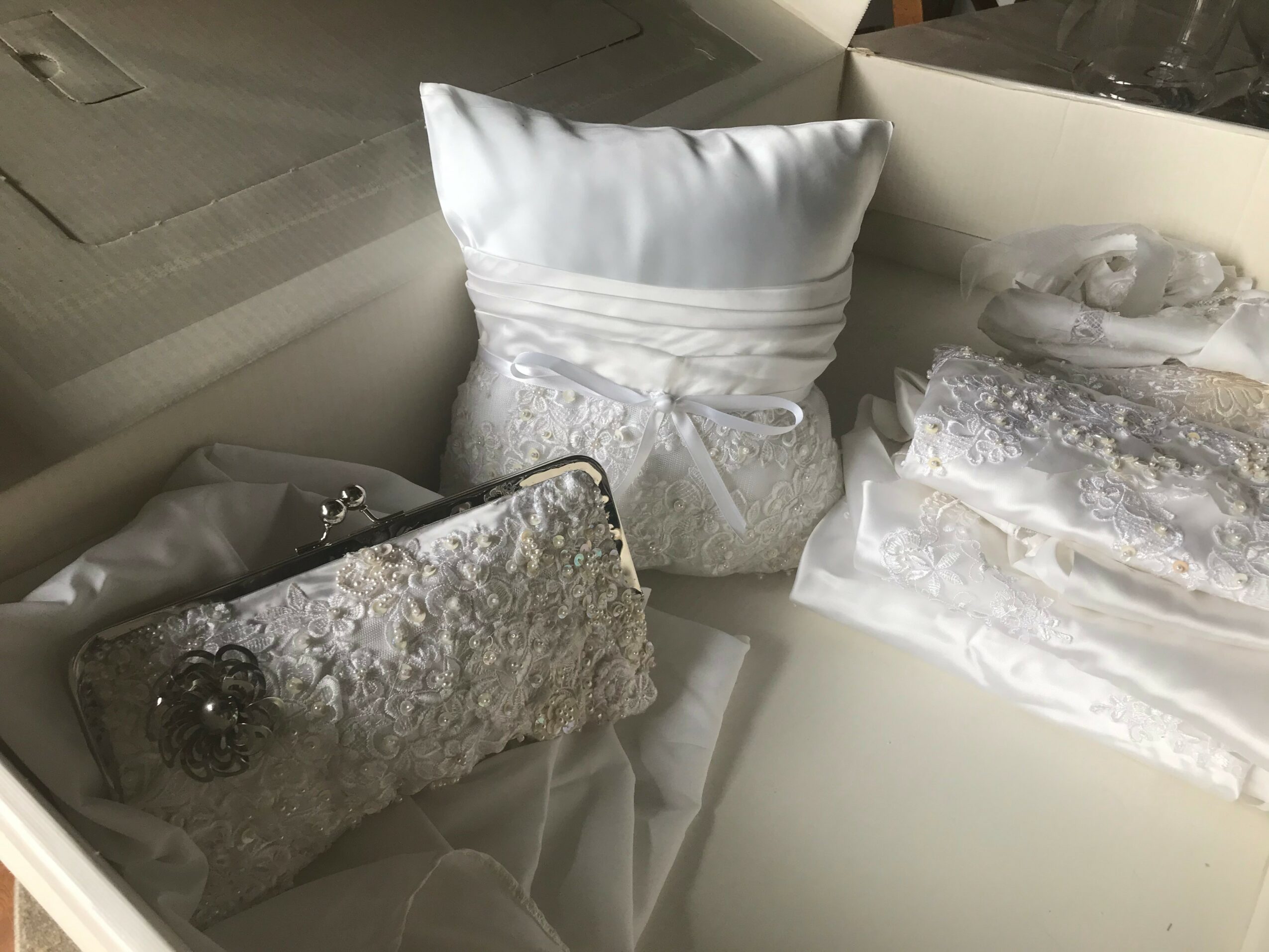 A white pillow with lace and ribbon details, a silver clutch with lace and a brooch, and a lace fabric piece inside a box.