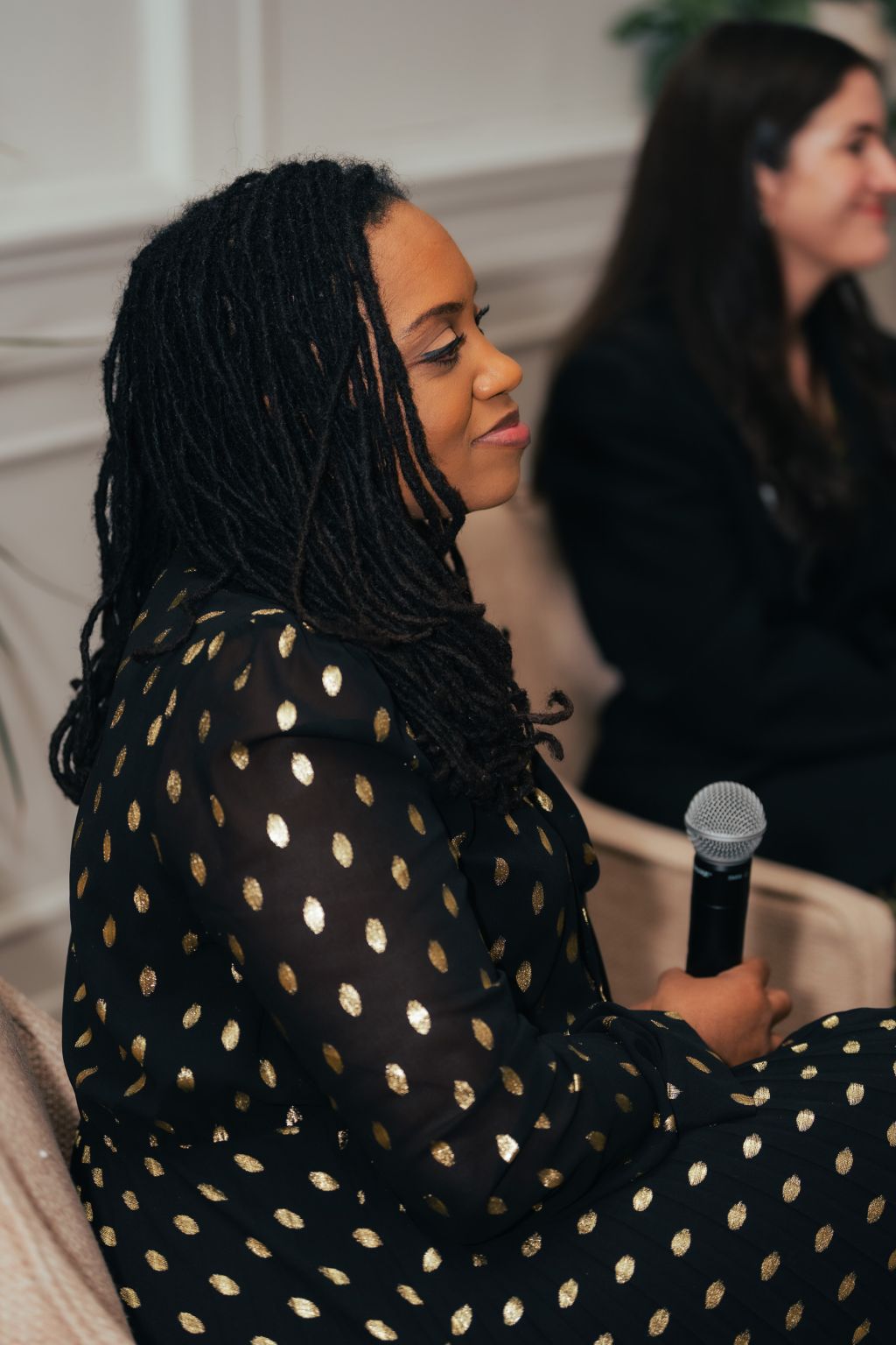 Woman with long braided hair holding a microphone, wearing a black dress with gold polka dots, sitting indoors.