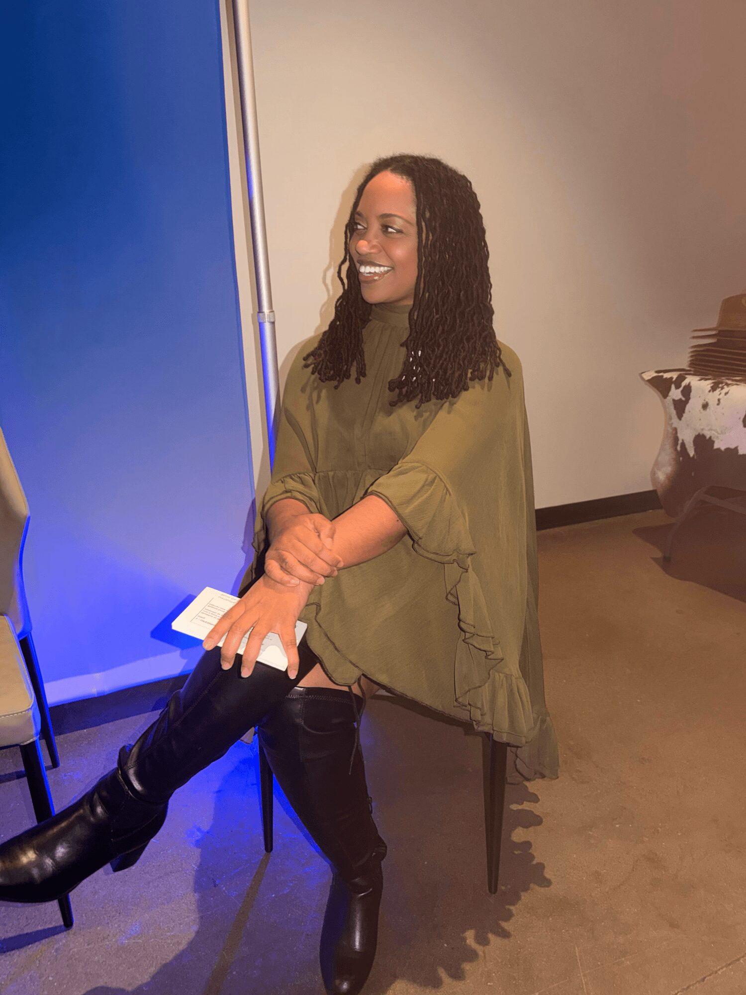 Woman with curly hair smiling, wearing a green top and black boots, sitting on a chair in an indoor setting.