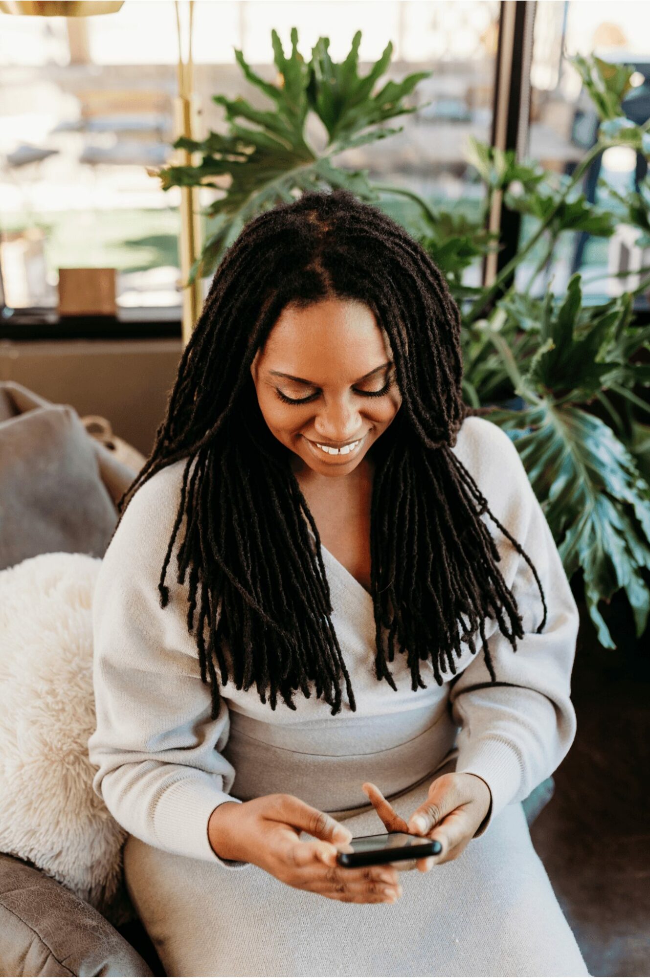 Woman with long dreadlocks smiling while looking at her phone in a bright indoor space.
