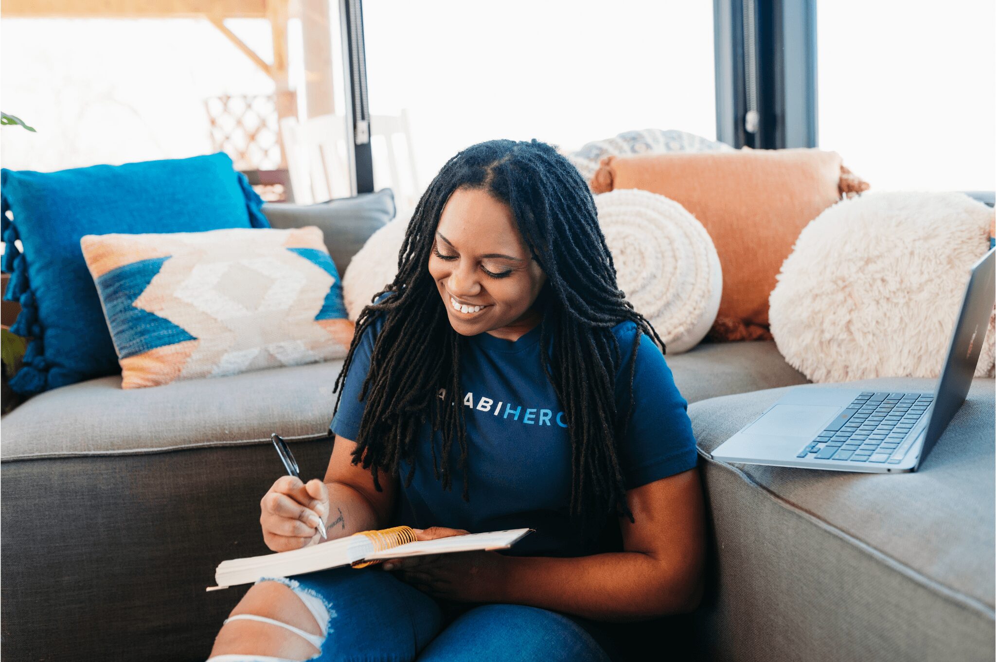 Woman sitting on a sofa, writing in a notebook, with colorful pillows and a laptop nearby, in a bright room.