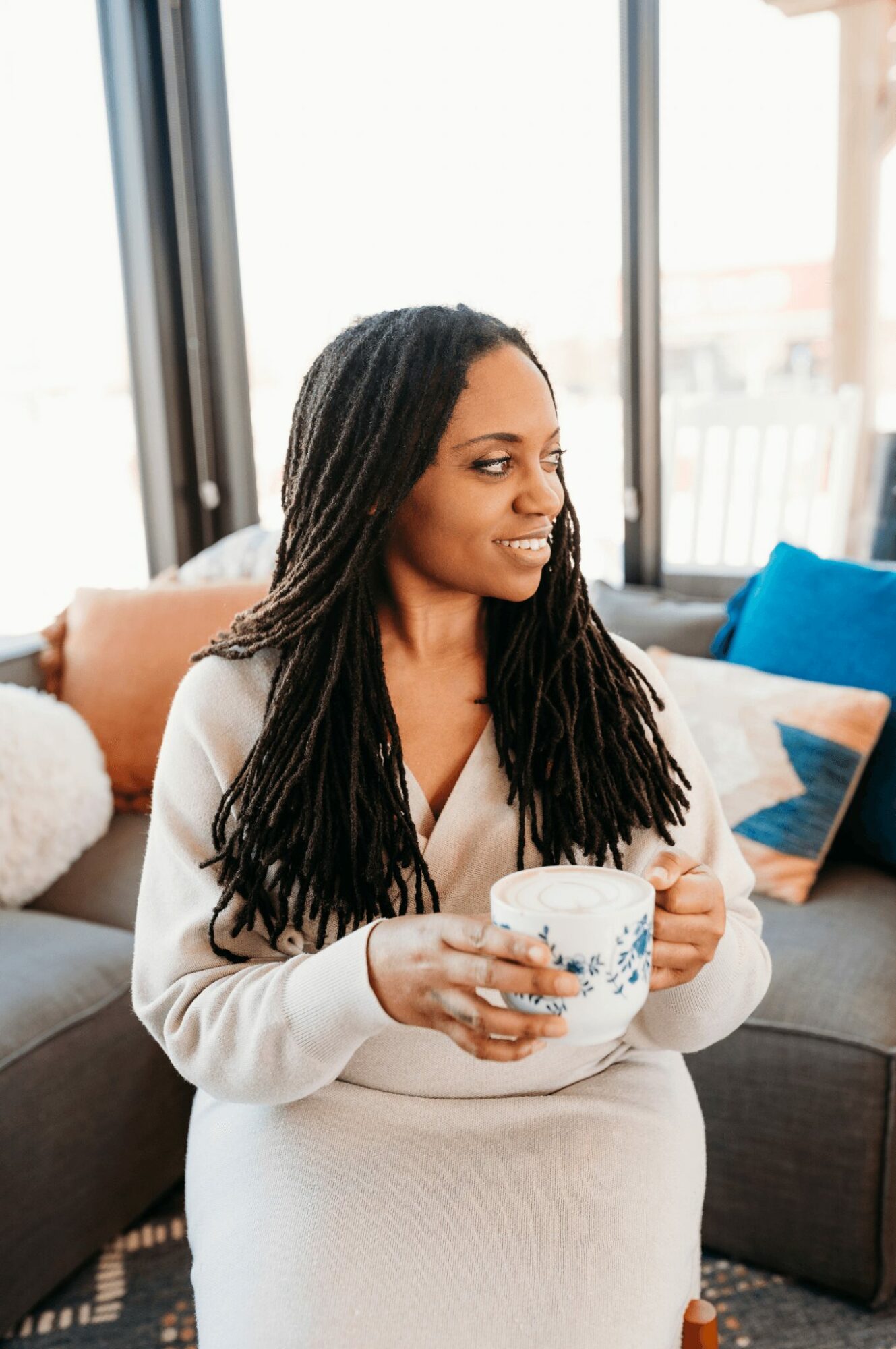 Woman with long dreadlocks sitting on a sofa, holding a mug, smiling, with large windows behind her.