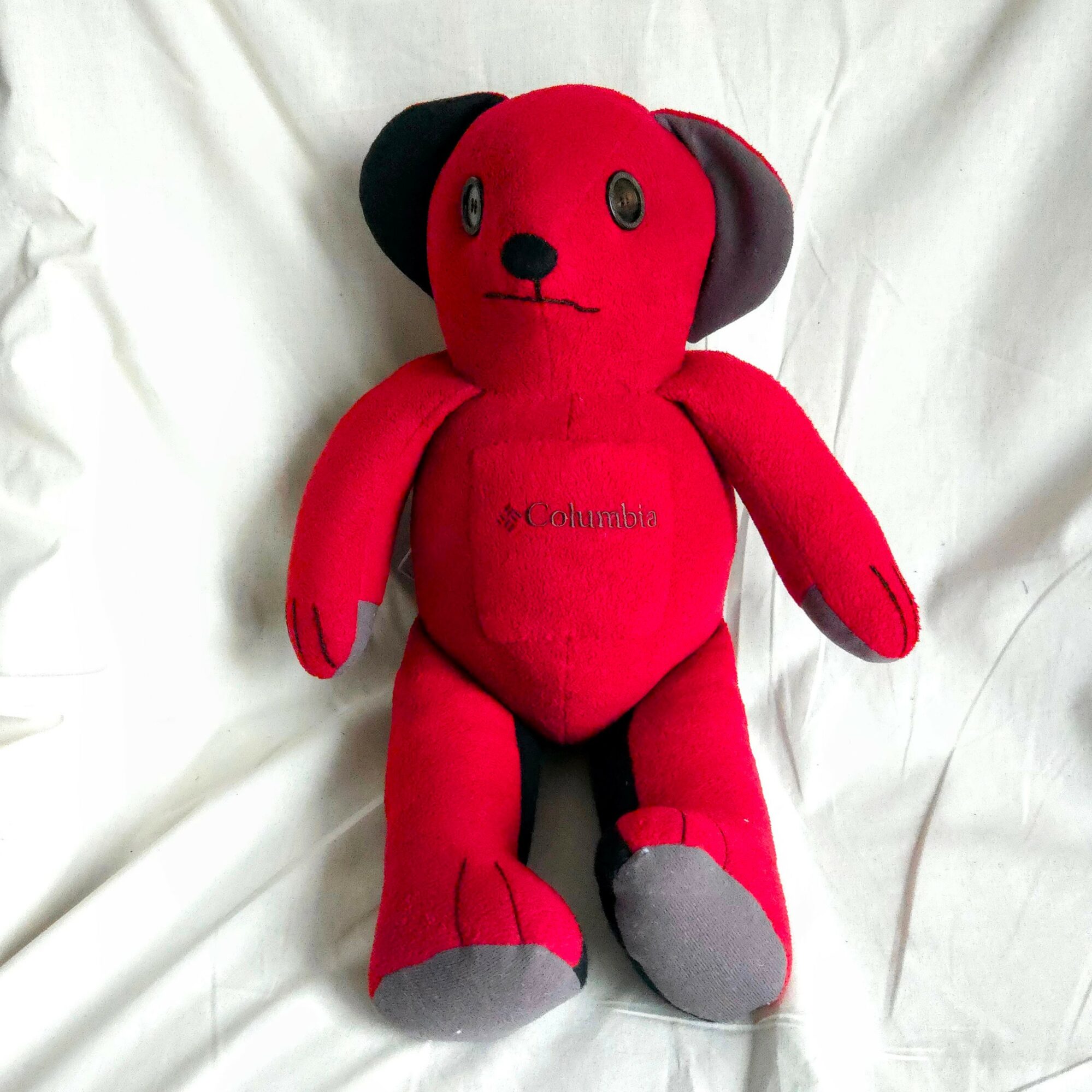 Red plush teddy bear with black ears and gray feet, lying on a white fabric surface.
