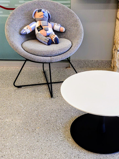 Gray upholstered chair with black metal legs, a stuffed robot toy on it, and a white round table with a black base nearby.