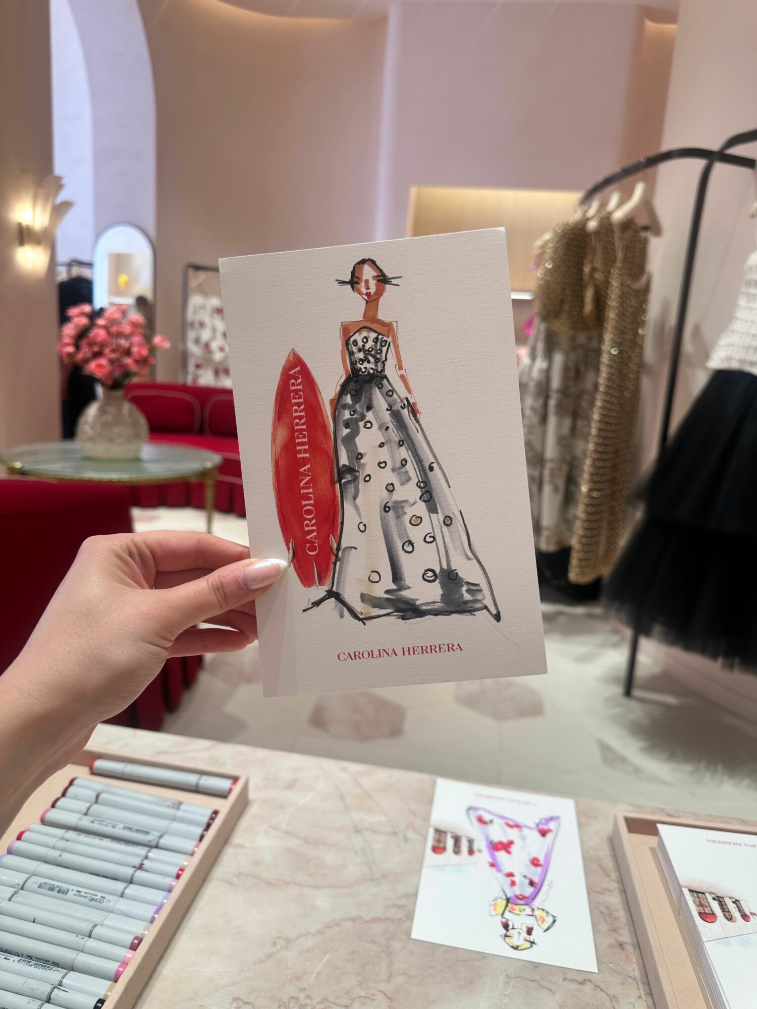 Hand holding a card with a fashion illustration of a woman in a polka dot dress and red sash, on a table.