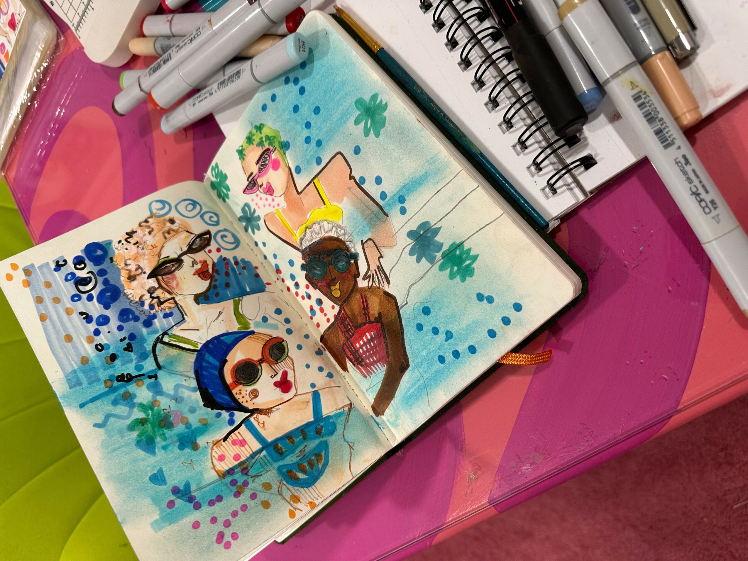 Open sketchbook with colorful illustrations of children, surrounded by markers and art supplies, on a pink surface.