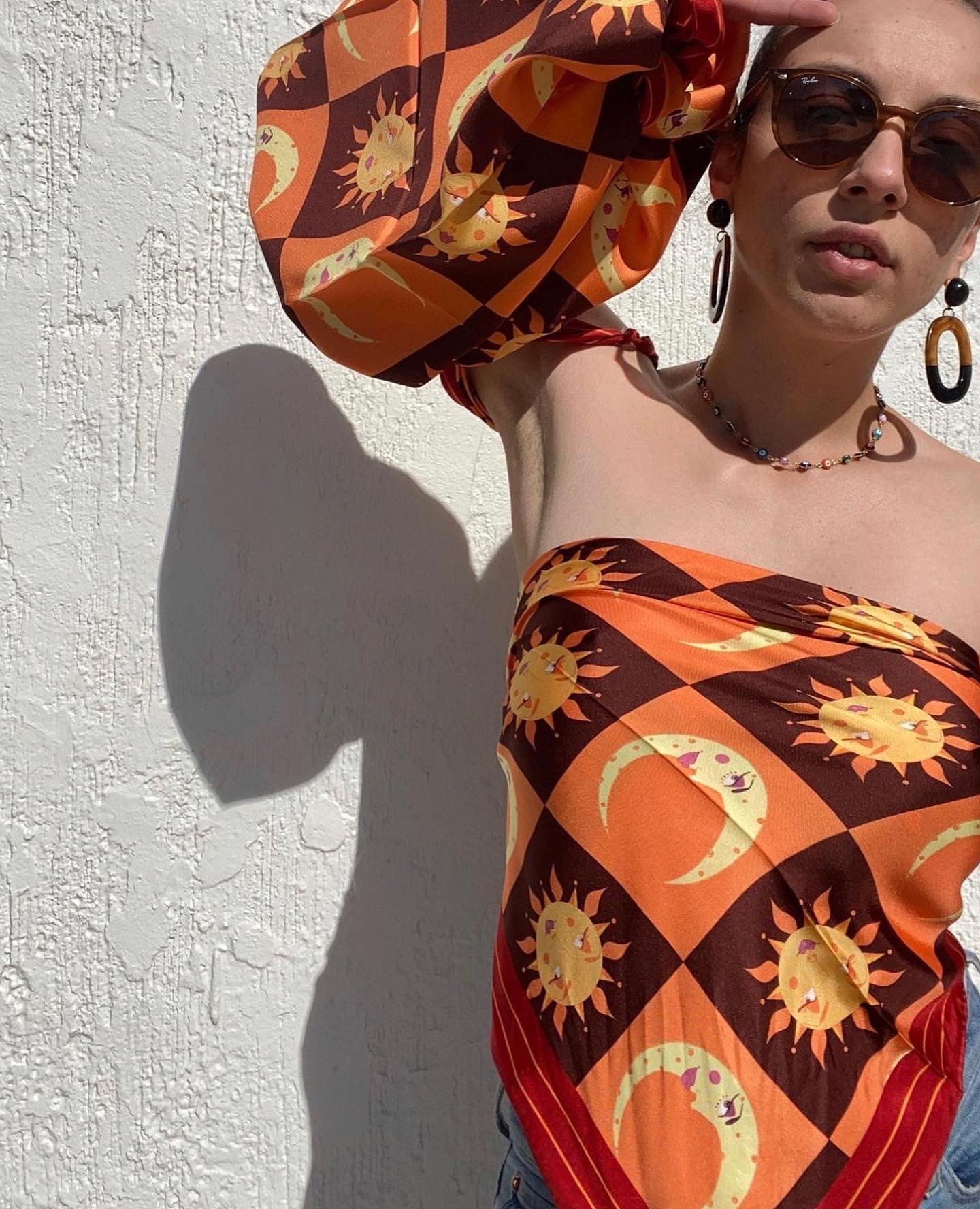 Woman wearing sunglasses and a patterned strapless top, standing against a white textured wall, shadow cast on wall.